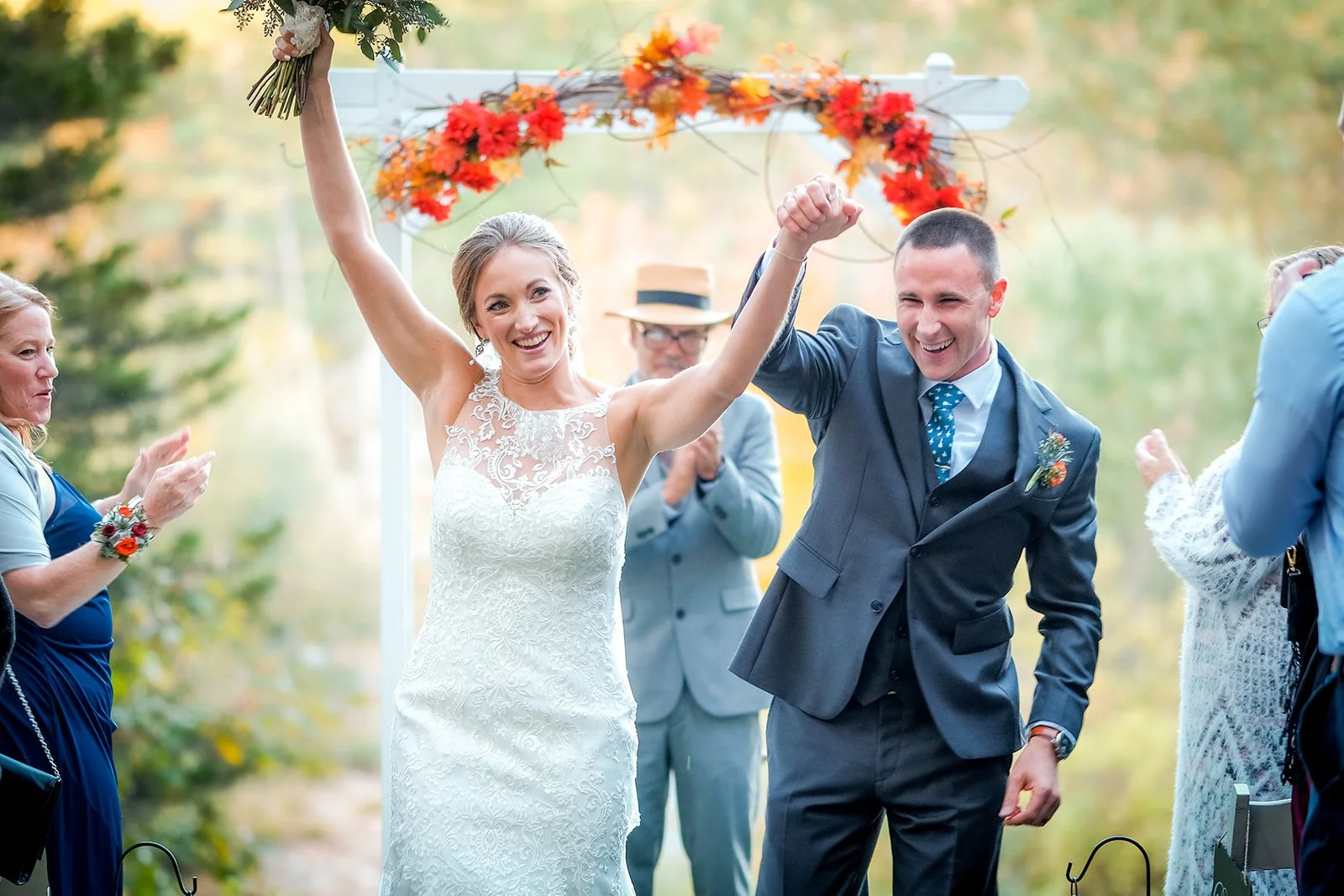 A bride and groom celebrating outdoors at their wedding, holding hands and smiling, surrounded by guests clapping and cheering, with a floral arch decorated with fall leaves in the background.