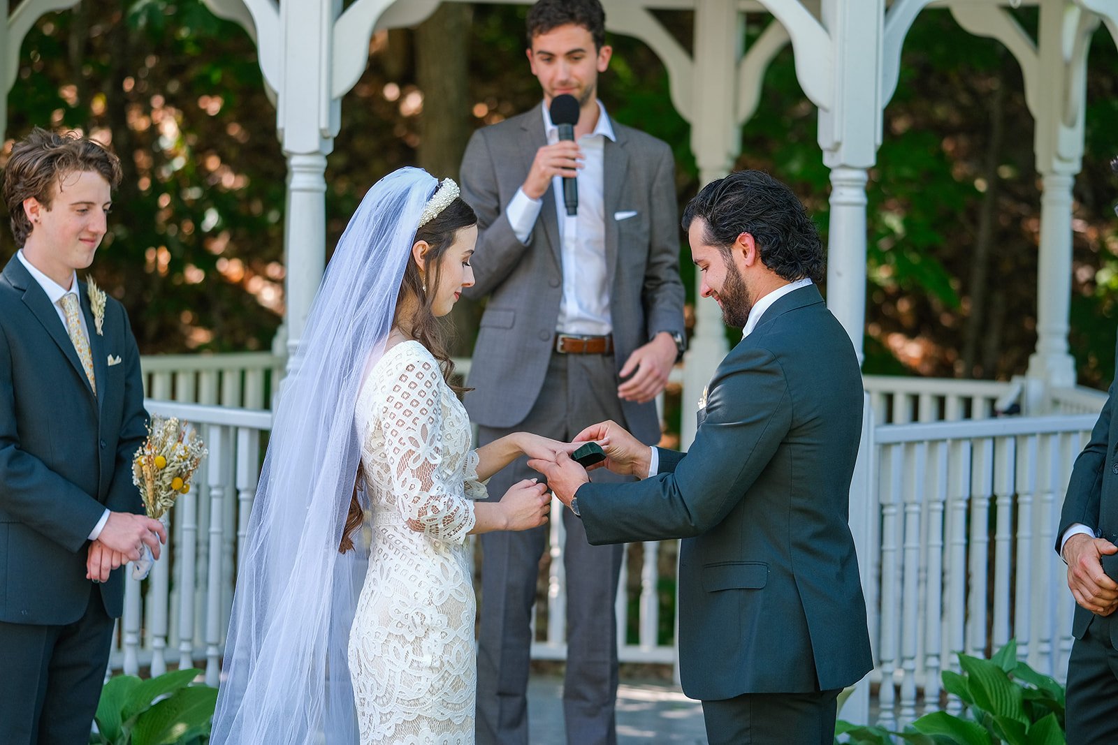 A couple exchanging wedding rings during an outdoor wedding ceremony under a white gazebo, with a man as the officiant standing behind them, flanked by a woman and a man in formal attire.
