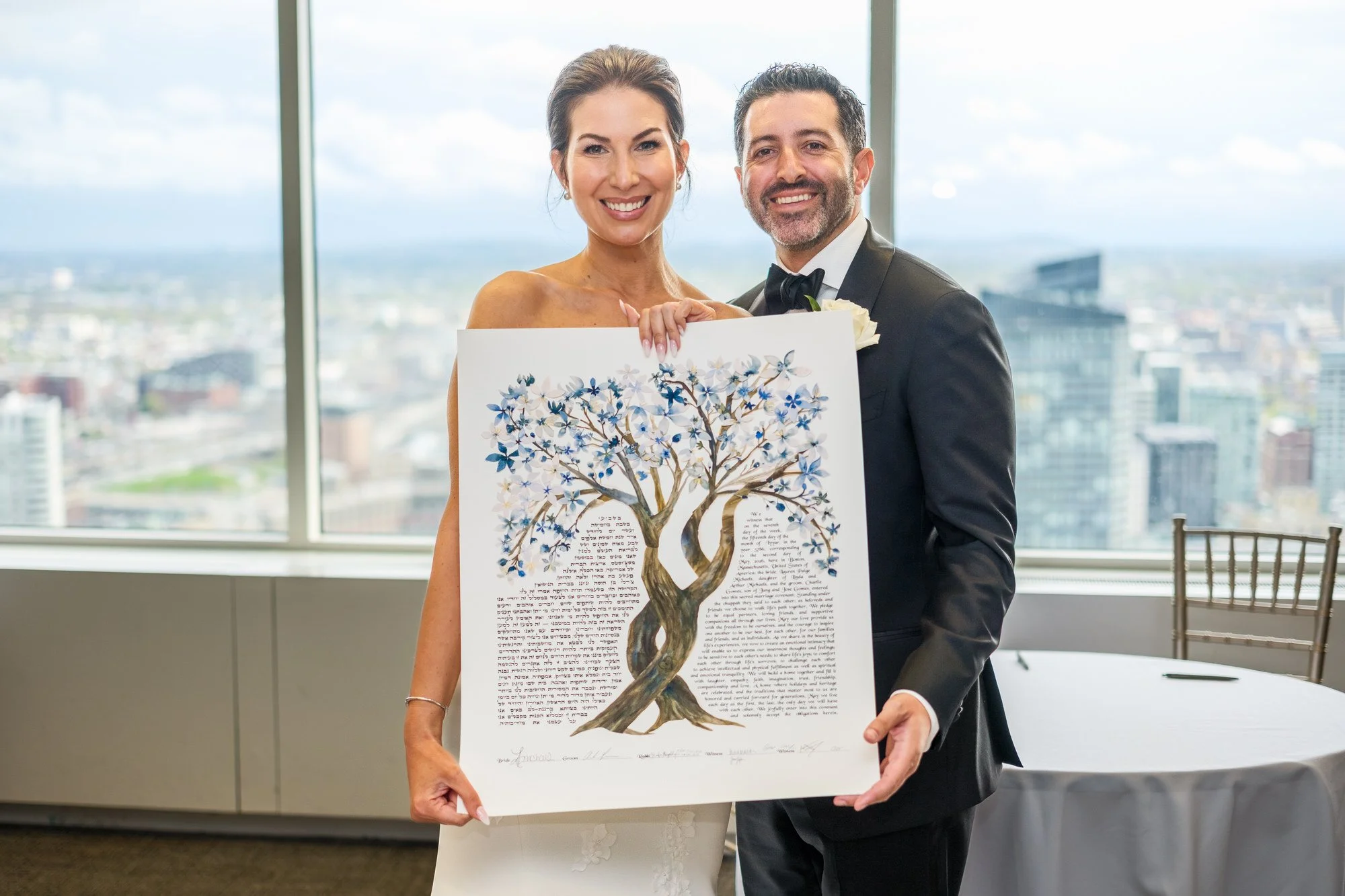 Bride and groom proudly displaying their signed ketubah with illustrated tree artwork and Boston skyline behind them