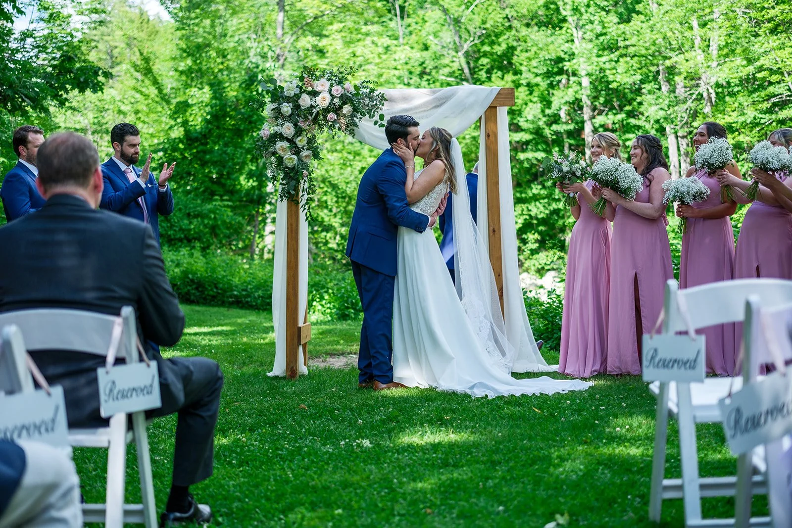 A couple sharing a kiss during a wedding ceremony outdoors, with a green forest background, officiant, bridesmaids in pink dresses holding bouquets, and chairs marked 'Reserved'.