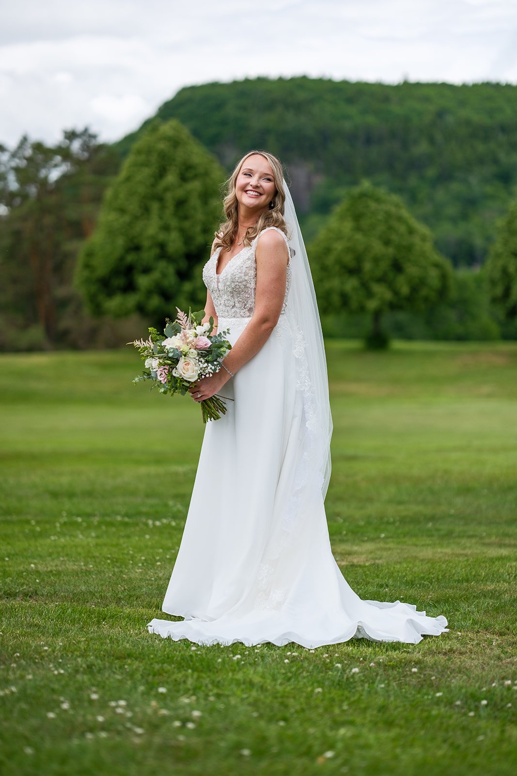 A bride in a white wedding dress holding a bouquet of flowers, standing on a green lawn with trees and a hill in the background.