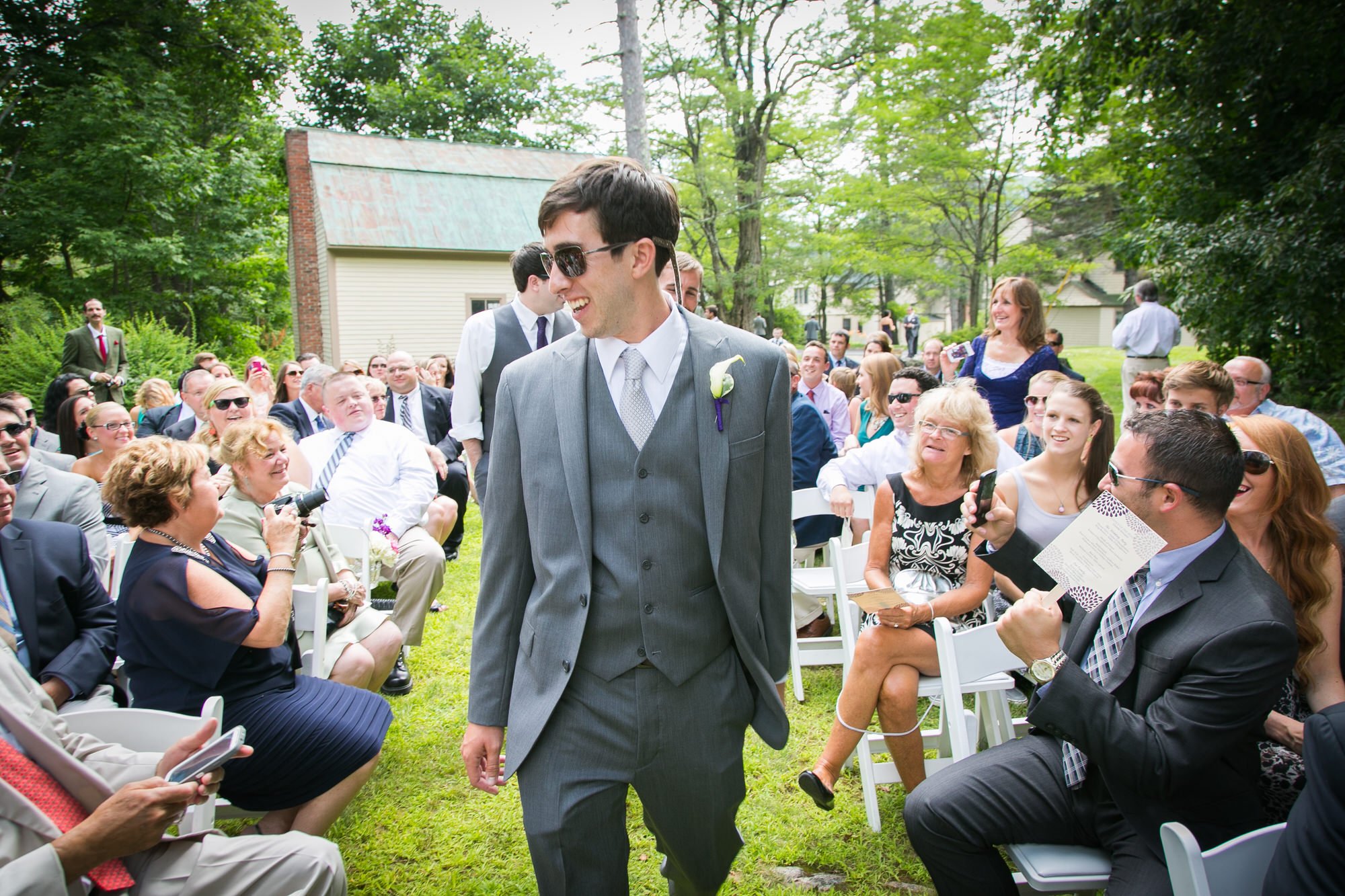 A man in a gray suit with sunglasses and a boutonniere is smiling while walking at an outdoor wedding ceremony. Guests seated in white chairs are watching and smiling, with some holding phones or cameras, in a lush green garden setting.