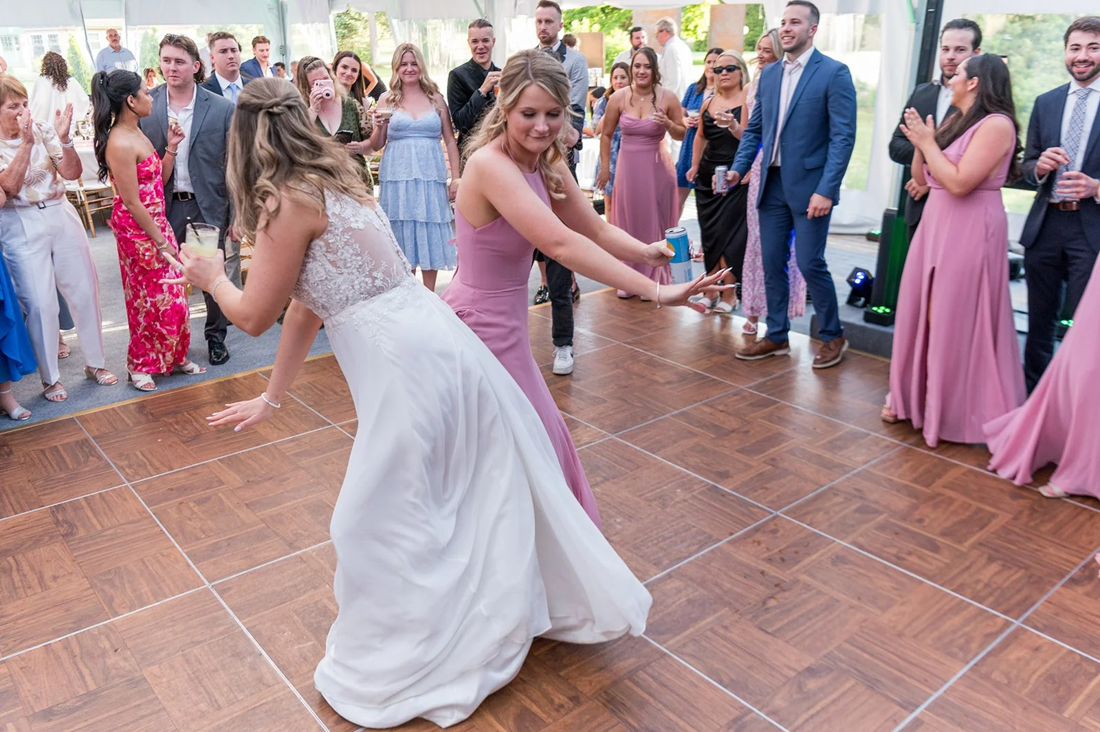 Wedding reception with guests dancing on a wooden floor, including a bride in a white wedding dress and a woman in a pink dress dancing together, surrounded by friends and family.