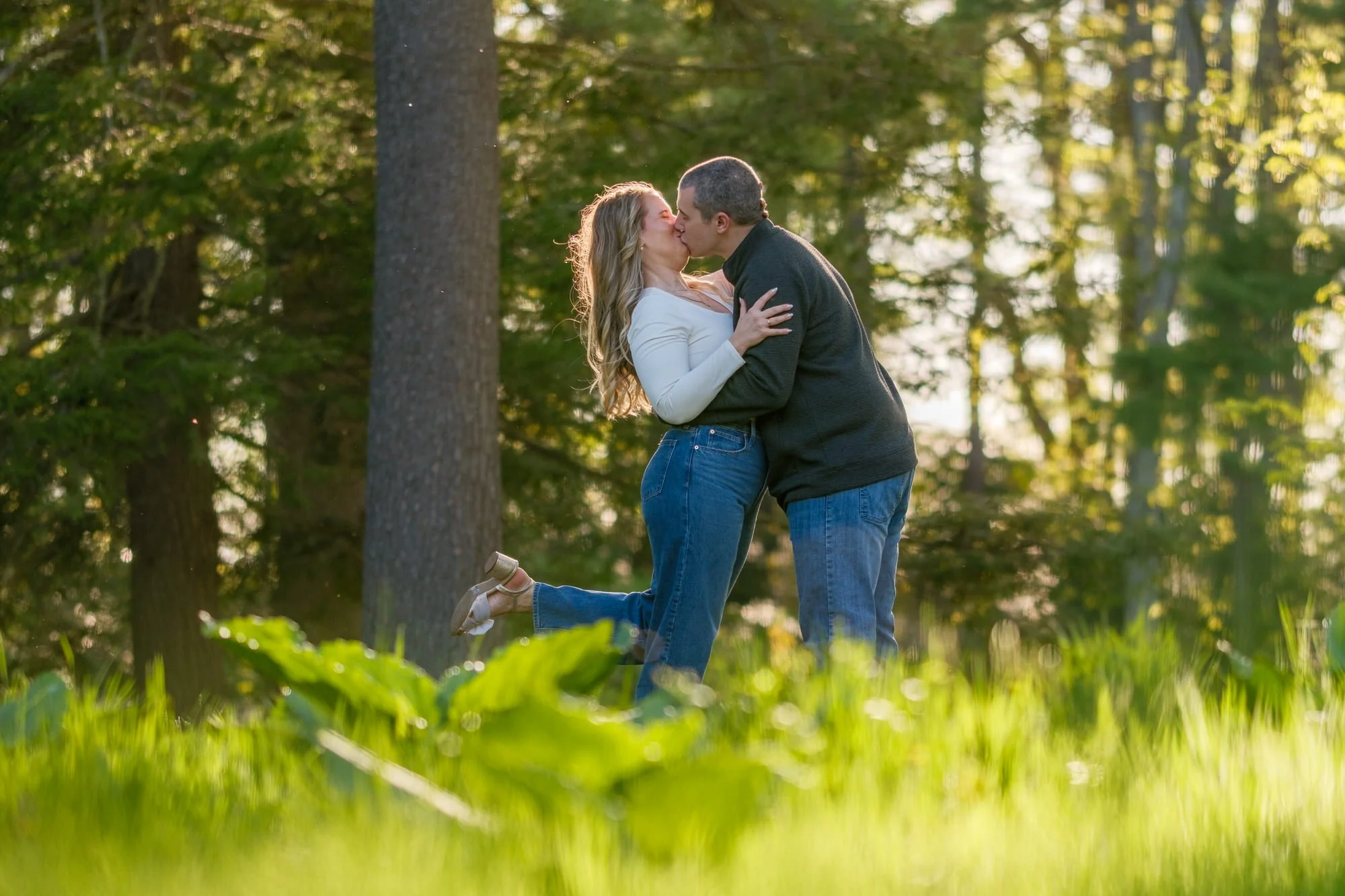Couple sharing a romantic kiss with her leg kicked up in a glowing pine forest at Maudslay State Park