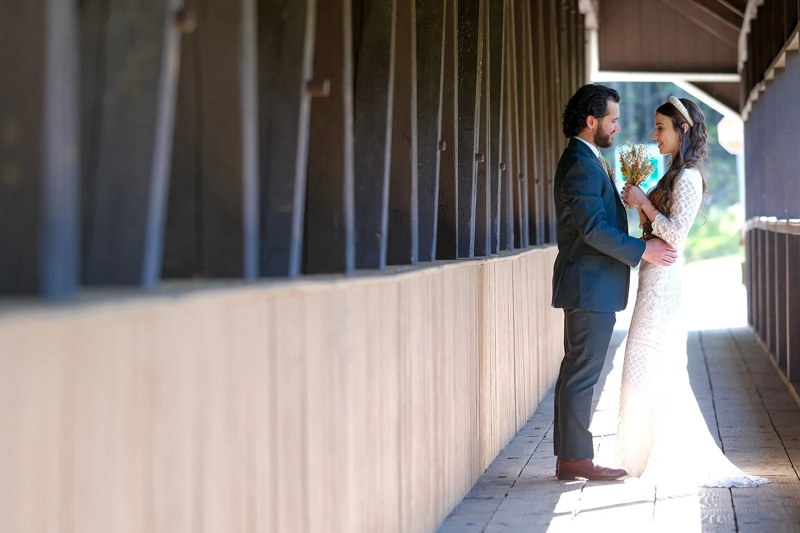 A bride and groom standing close together, holding hands and facing each other, inside a wooden covered walkway with sunlight in the background. The bride is wearing a white lace wedding dress and holding a bouquet, the groom is in a dark suit.