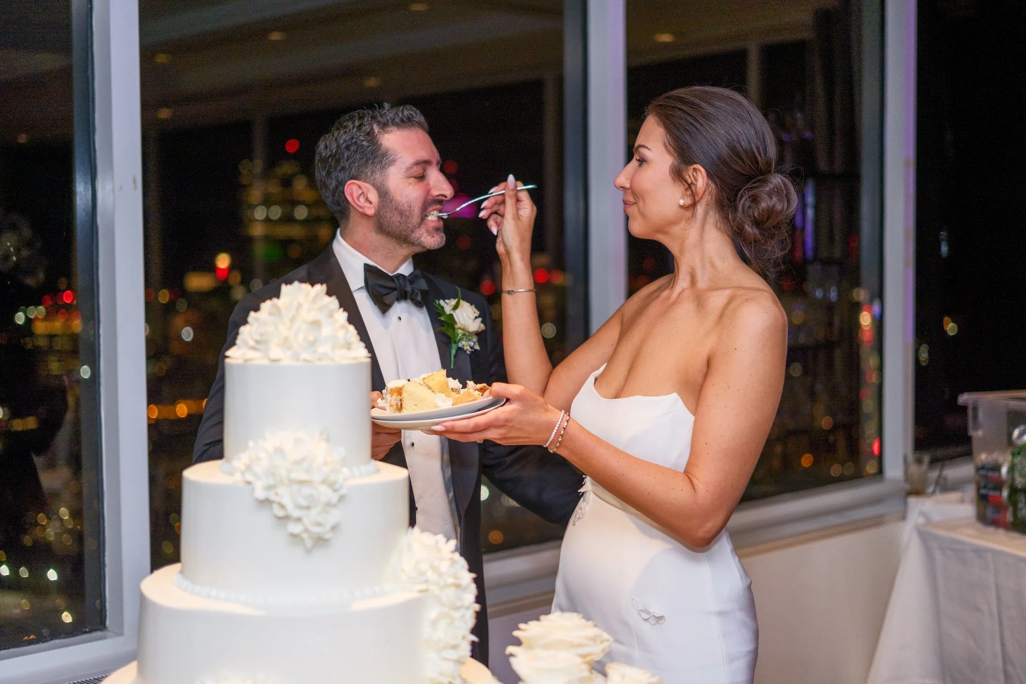 Bride playfully feeding wedding cake to groom at the UMass Club Boston reception with Boston night skyline