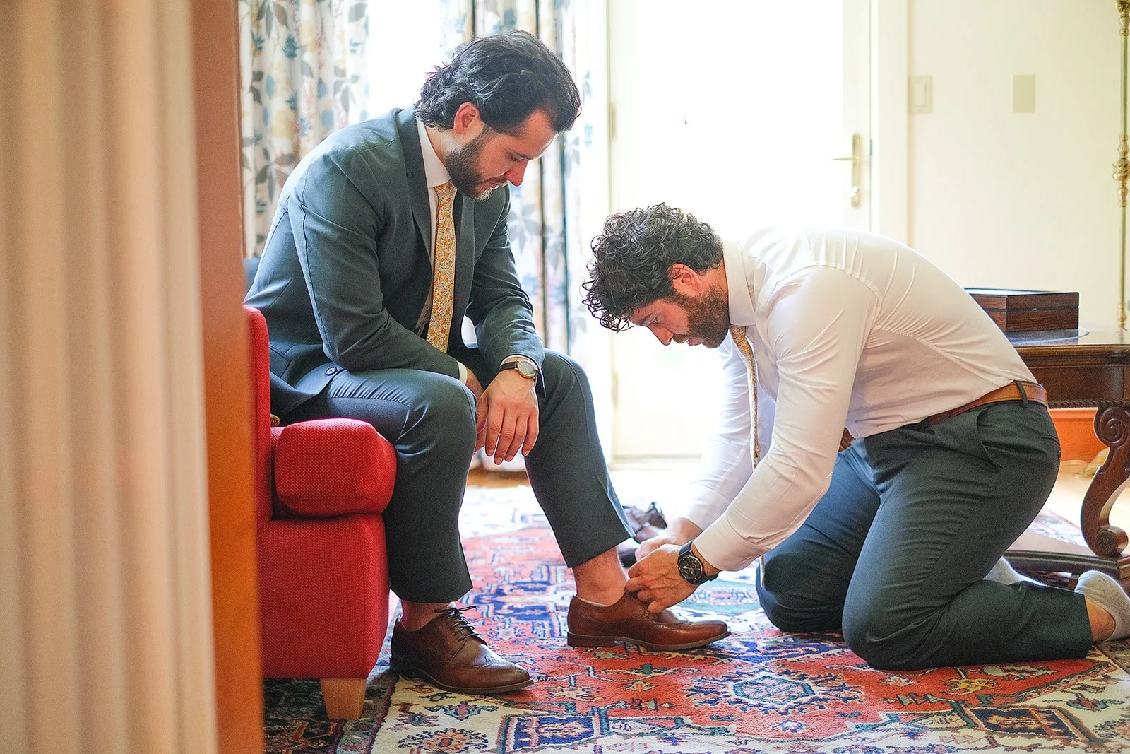 Two men dressed in suits, one seated on a red sofa and the other kneeling on a patterned rug, helping the seated man put on a brown dress shoe in a well-lit room.