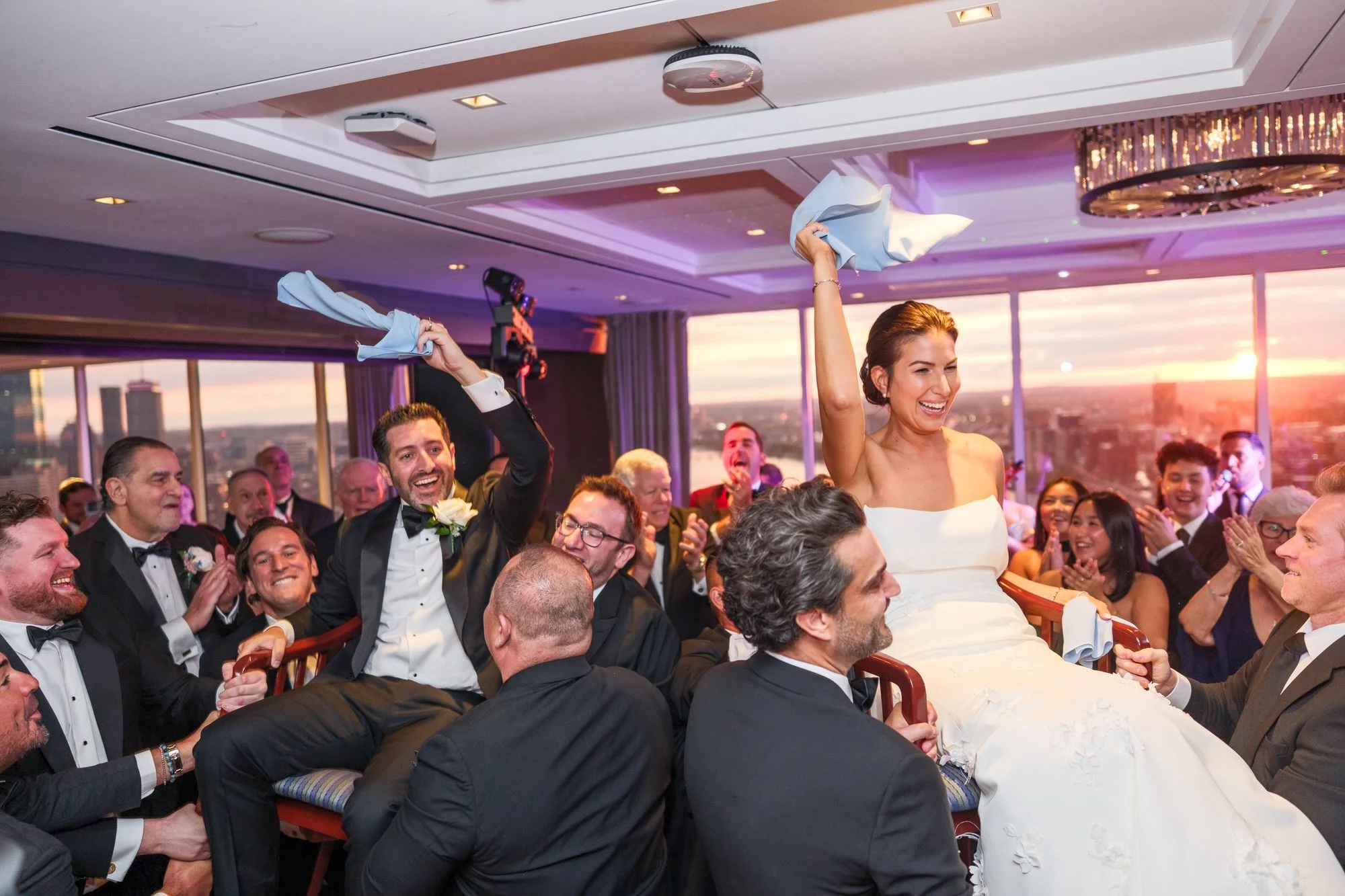 Bride lifted high in a chair during the traditional Jewish chair dance (hora) at the UMass Club Boston reception at sunset