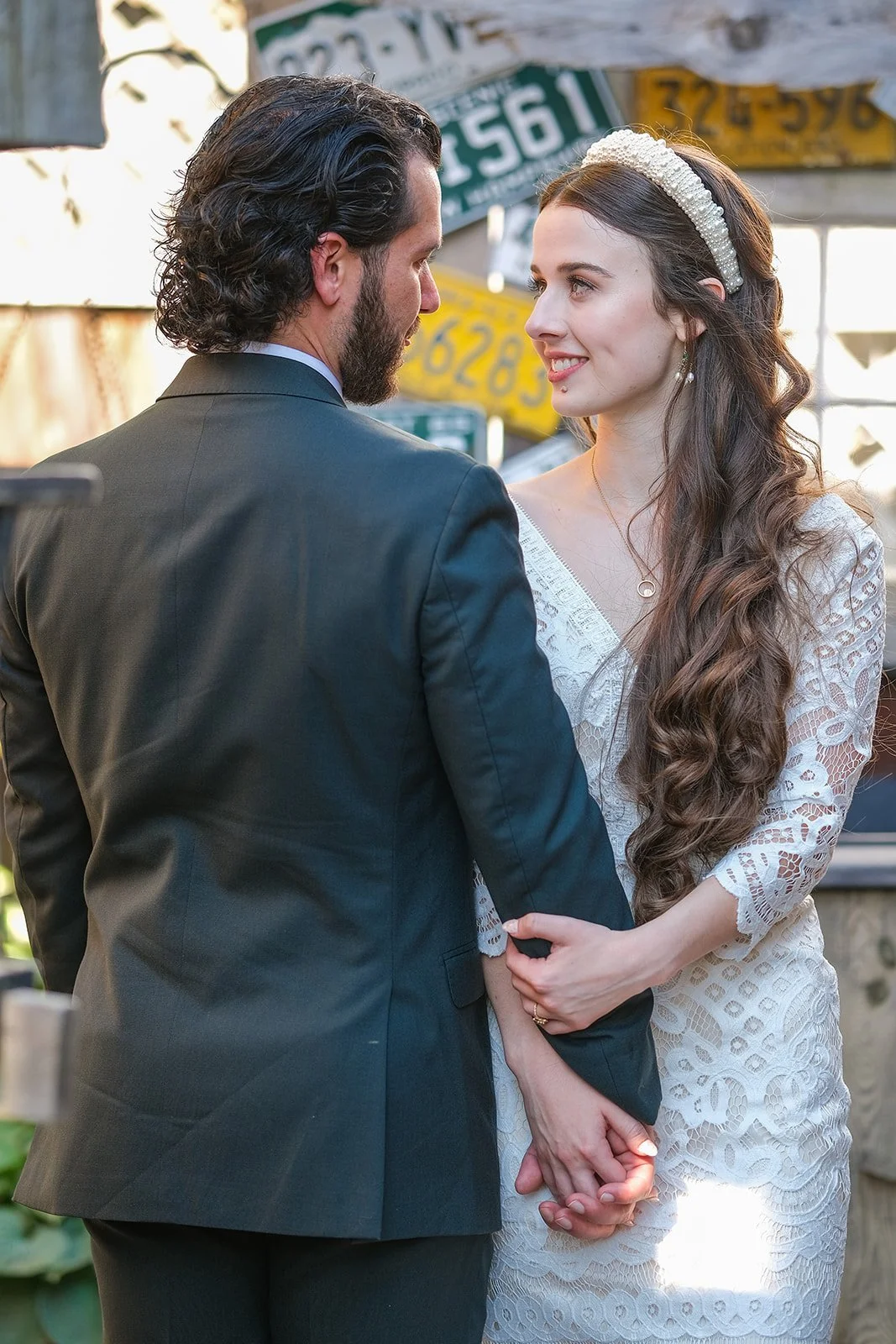 A man and woman hold hands and look at each other in an outdoor setting with vintage license plates in the background.