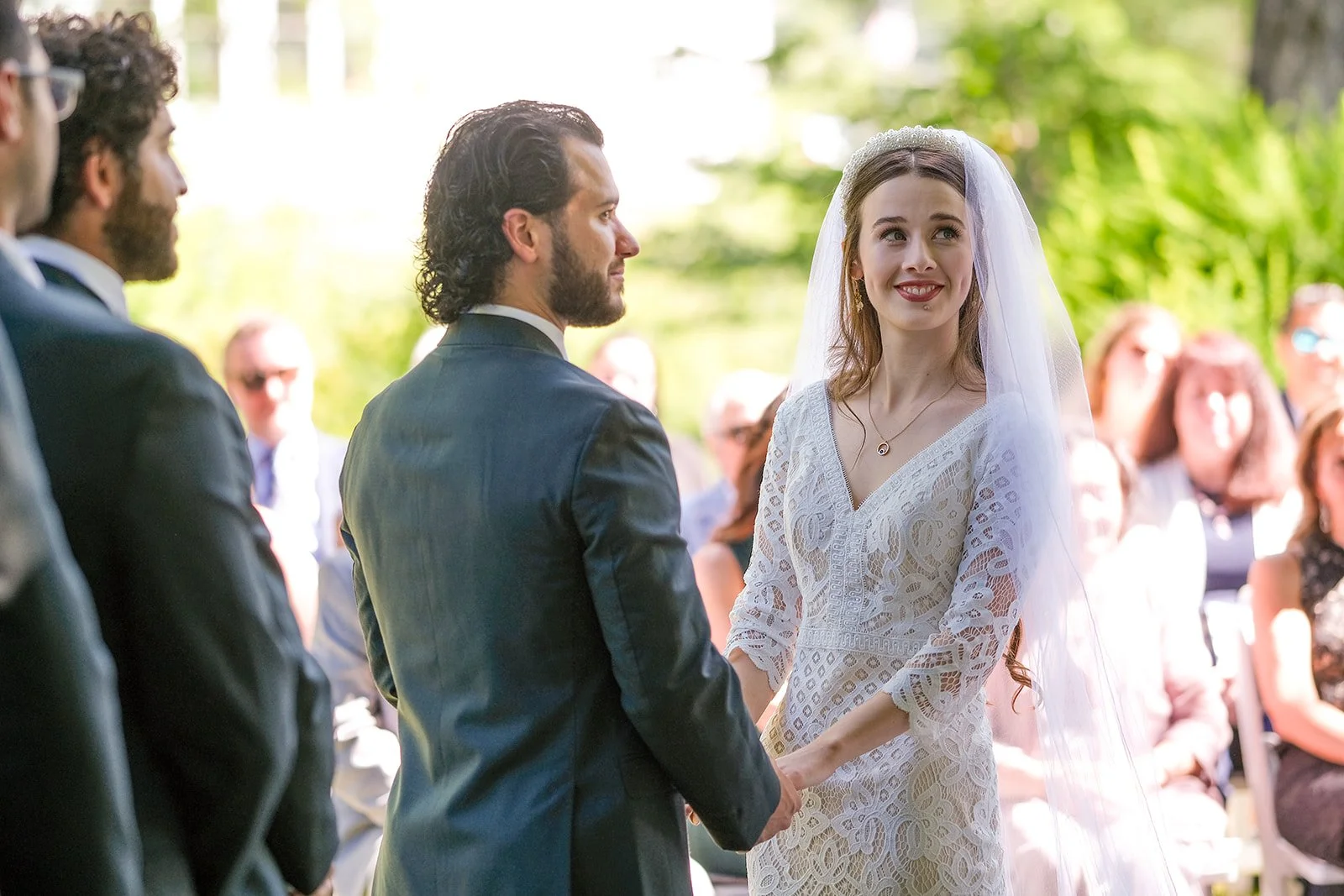 A bride and groom exchanging vows at an outdoor wedding ceremony, with guests seated behind them. The bride is smiling and wearing a white lace dress and veil, while the groom is in a dark suit. The setting is lush and green.