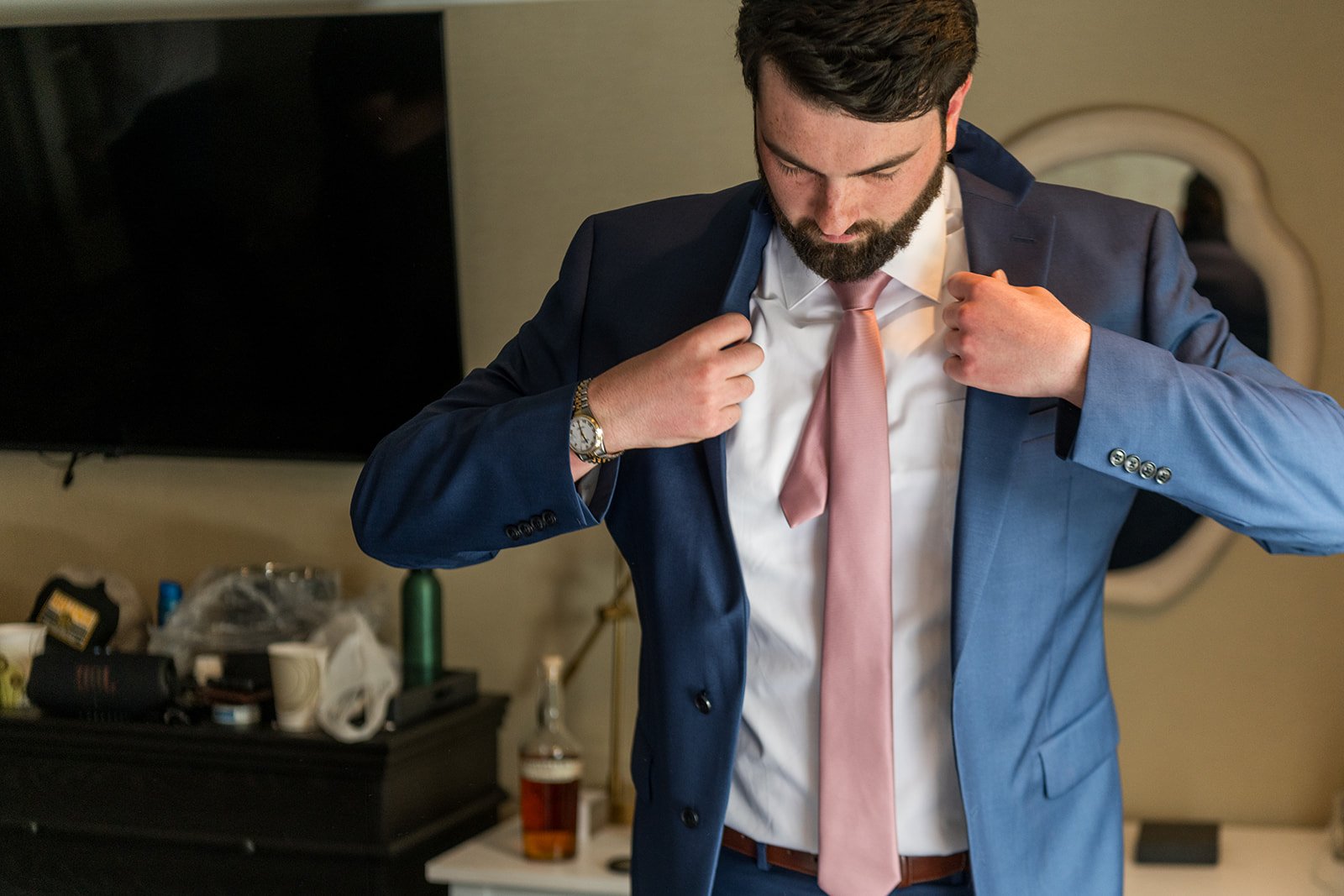 Man in a navy suit adjusting a pink tie in a hotel room.