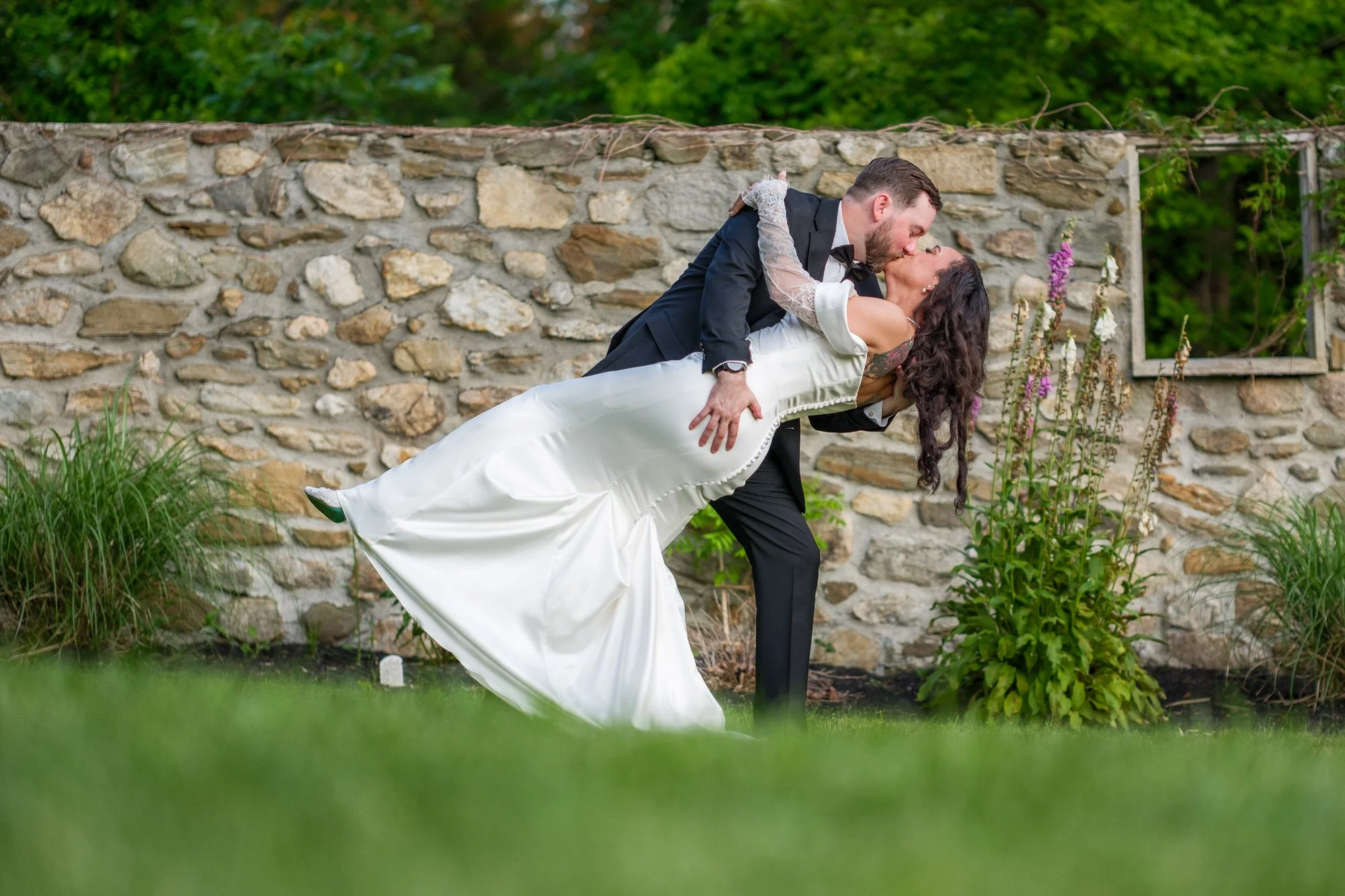 Groom dipping bride during portraits by stone wall at Hyla Brook Estate