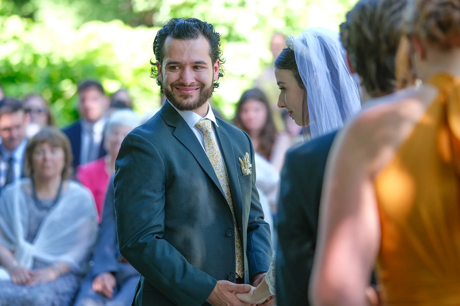 A groom with dark, curly hair and a beard in a blue suit holding hands with a bride with dark hair and a veil at an outdoor wedding ceremony. Guests are seated in the background.