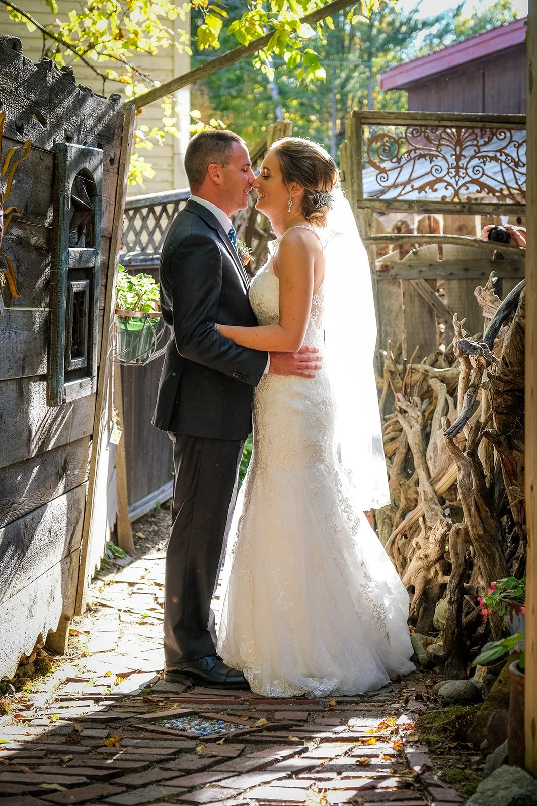 A newlywed couple standing close, embracing and about to kiss in a sunlit outdoor setting surrounded by rustic wooden structures and greenery.