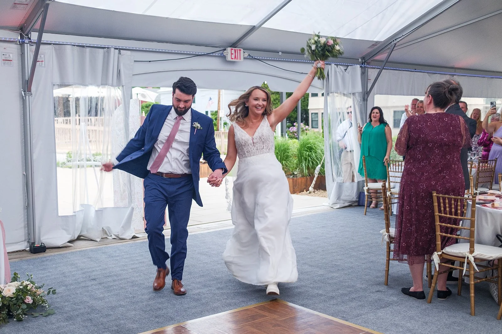 A bride and groom celebrating their wedding in a tent, holding hands and smiling, with guests standing and clapping around them.
