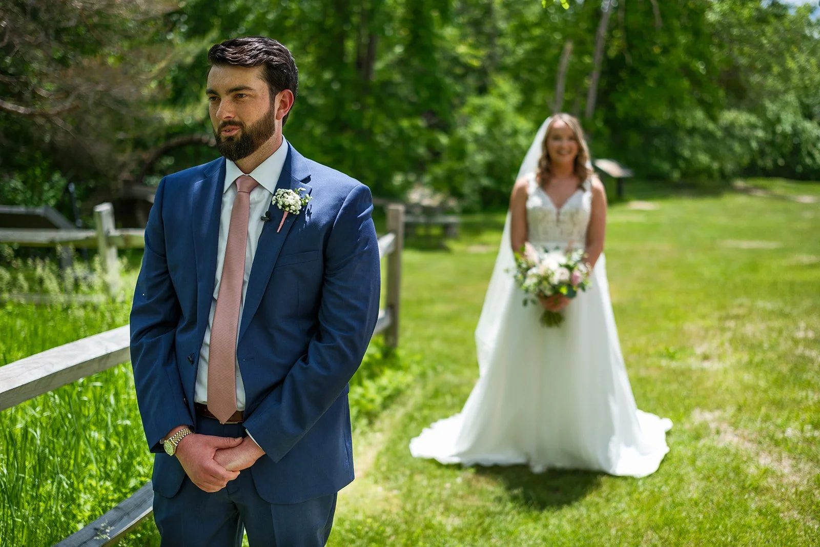 A groom in a blue suit and pink tie, standing outdoors on a grassy area and looking away, with a bride in a white wedding dress and veil holding a bouquet of flowers in the background, smiling.