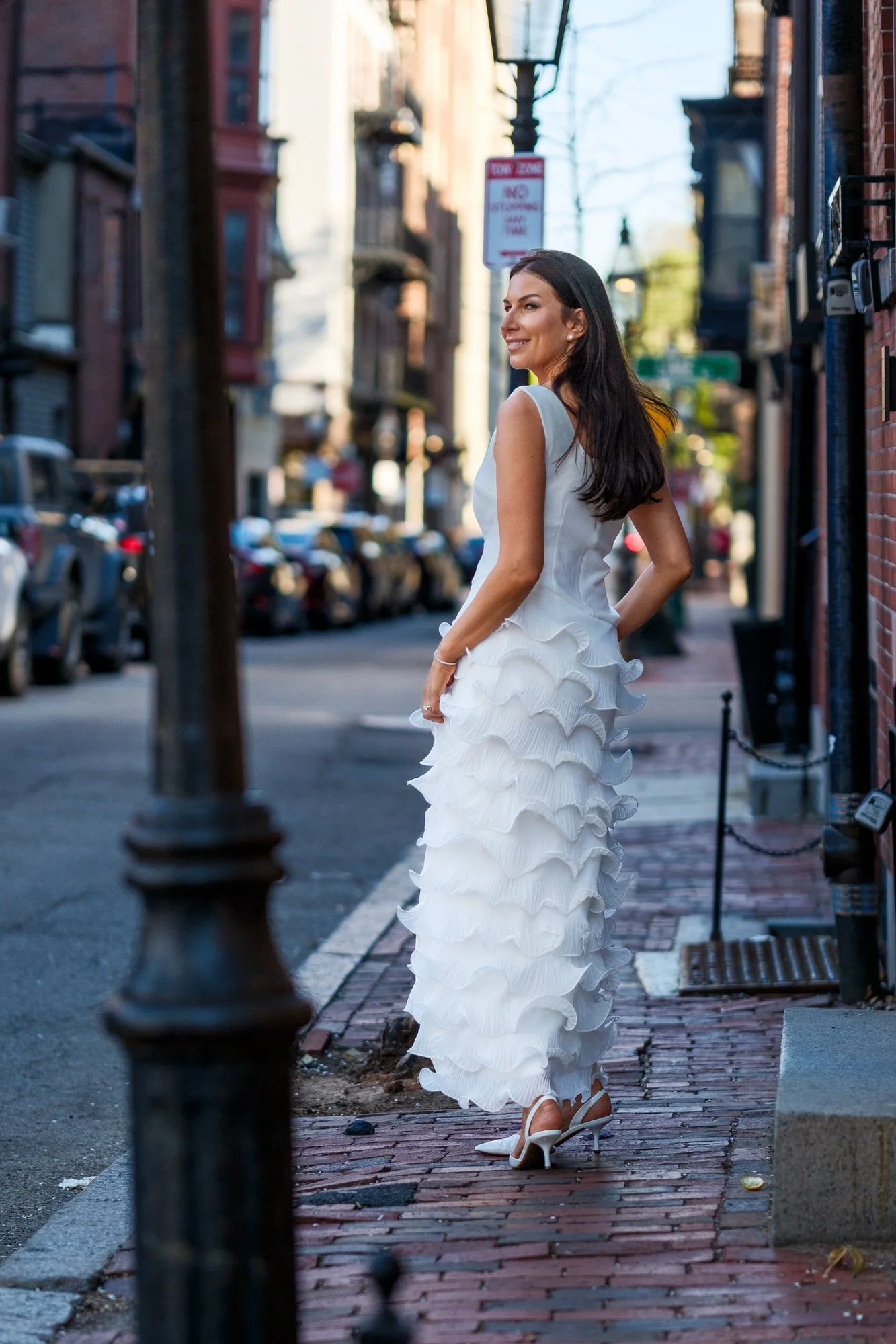 Bride Lauren glancing back over her shoulder on a brick sidewalk in Beacon Hill, her white tiered ruffle gown catching golden afternoon light as the historic neighborhood street stretches behind her