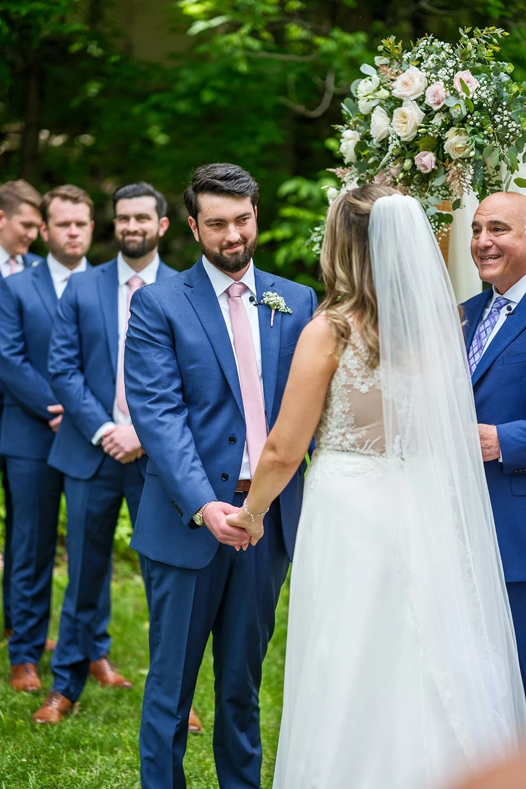 A wedding ceremony outdoors with a bride and groom holding hands, surrounded by groomsmen in blue suits, with a floral arrangement overhead and green trees in the background.