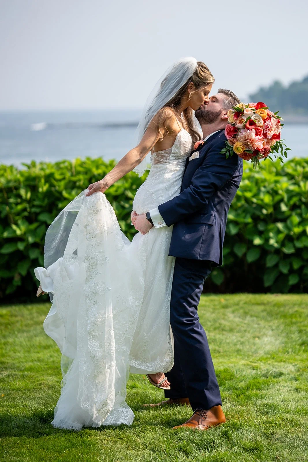Groom lifts bride for a romantic kiss with the York Maine coastline and Atlantic Ocean in the background