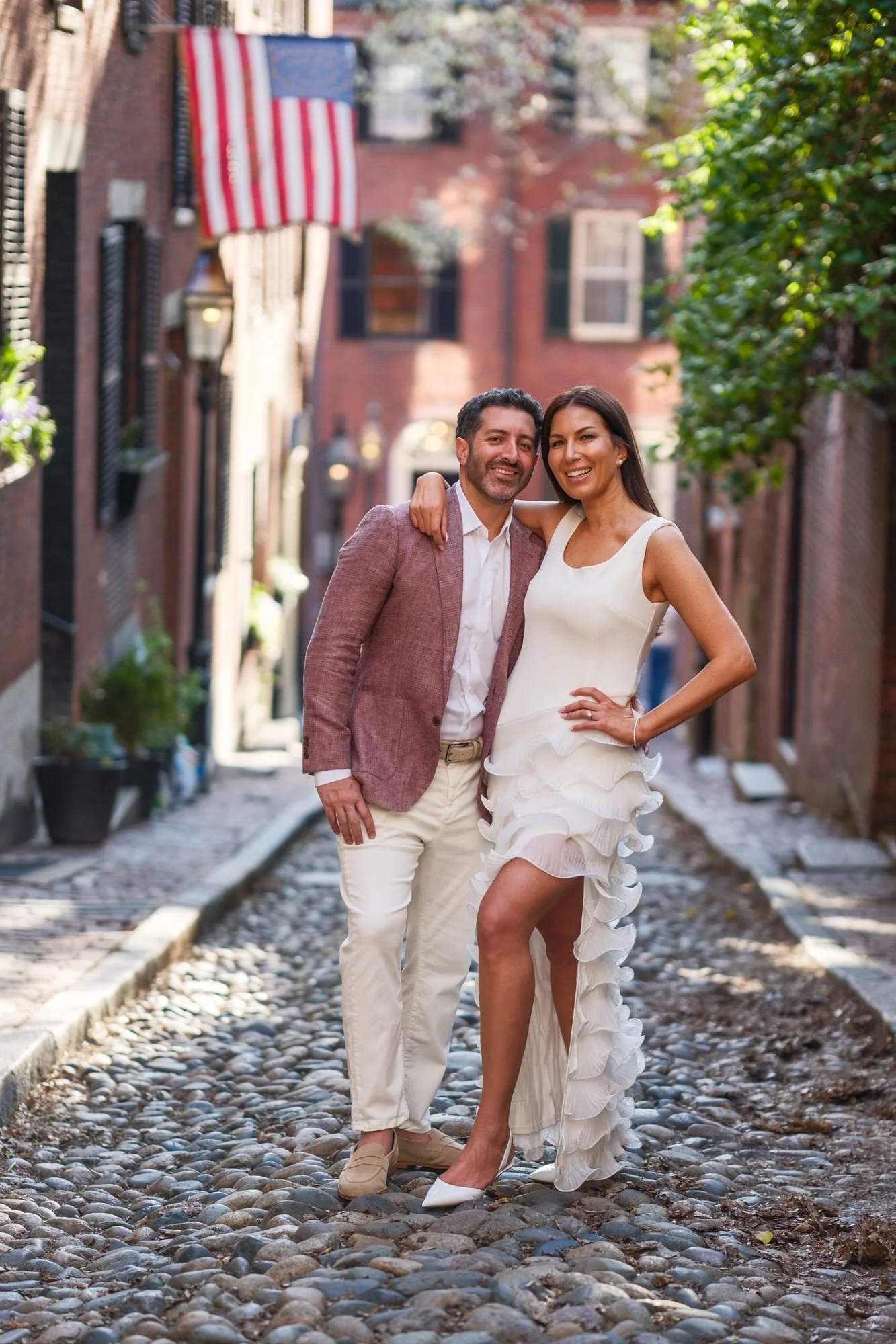 Lauren and Charlie posing together on the cobblestones of Acorn Street in Boston's Beacon Hill neighborhood, smiling with historic brick row houses and an American flag in the background