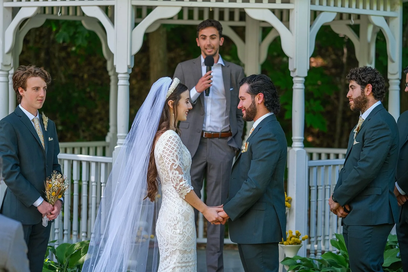 A couple getting married outdoors, holding hands and facing each other, with an officiant standing behind them. The bride wears a white lace dress and veil, and the groom wears a dark suit. Three groomsmen, one holding a bouquet, stand nearby. The se