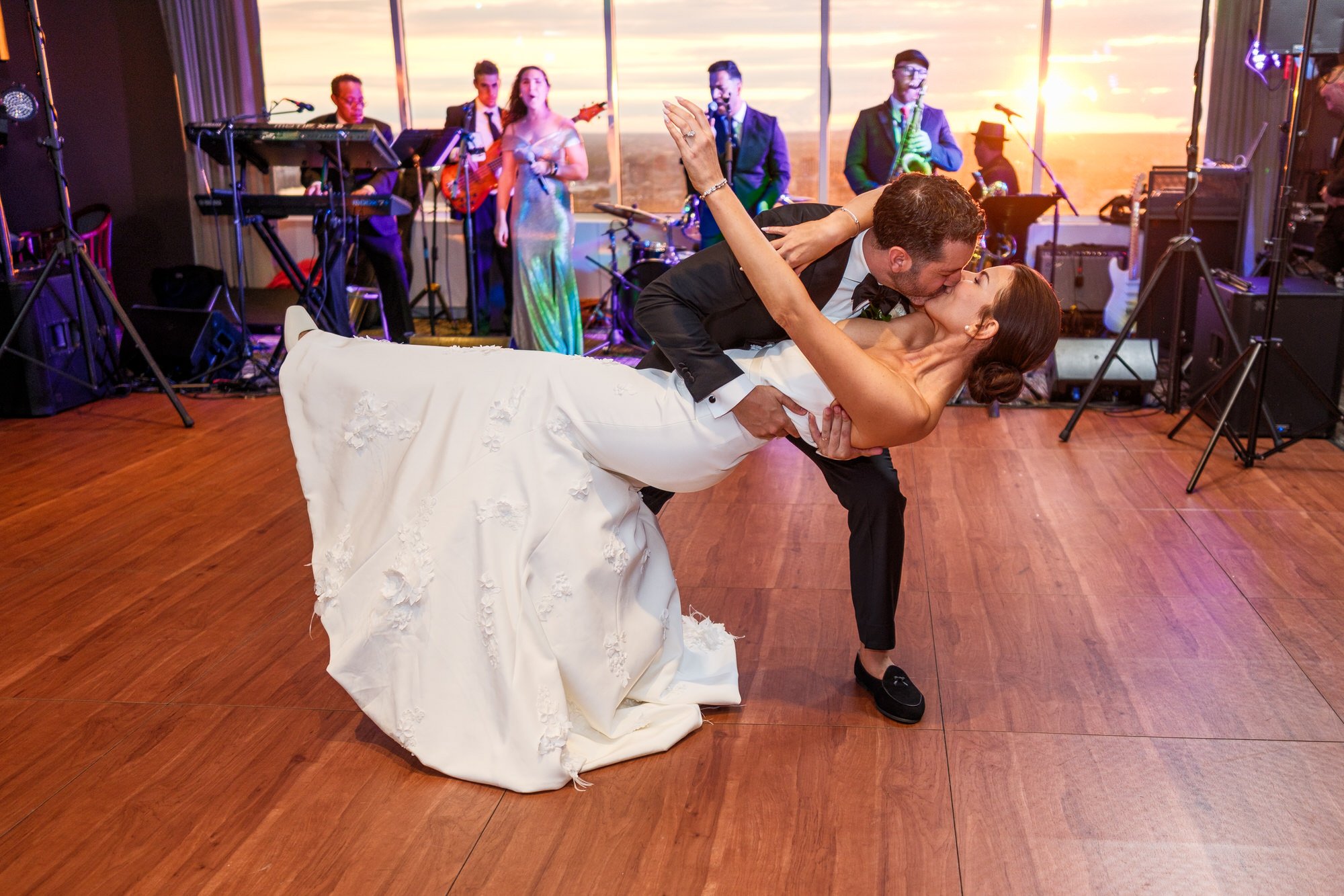 Groom dipping bride for a kiss during sunset first dance at the UMass Club Boston with live band performing behind them