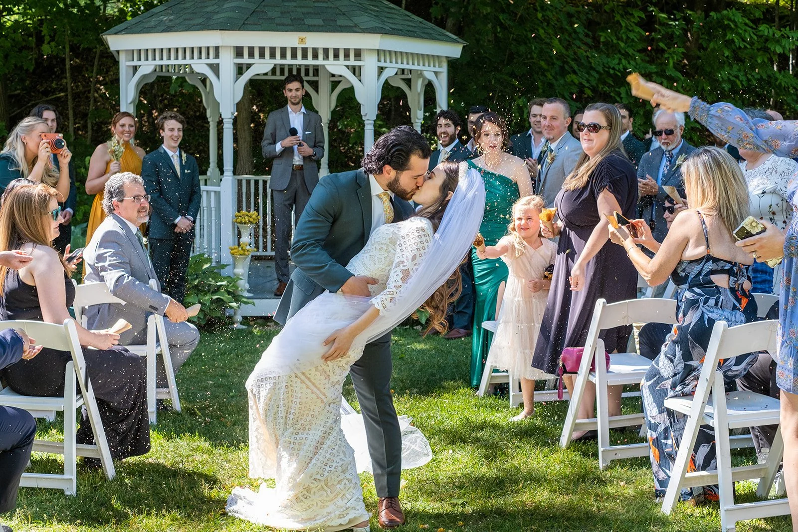 The bride and groom kiss during their outdoor wedding ceremony as guests watch and celebrate.