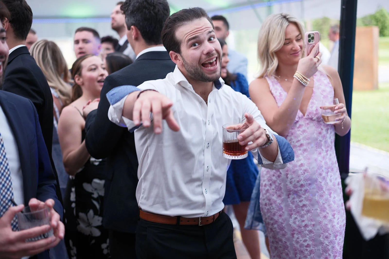 A group of people at a party, dancing and socializing, with one man in the center smiling and holding a drink.