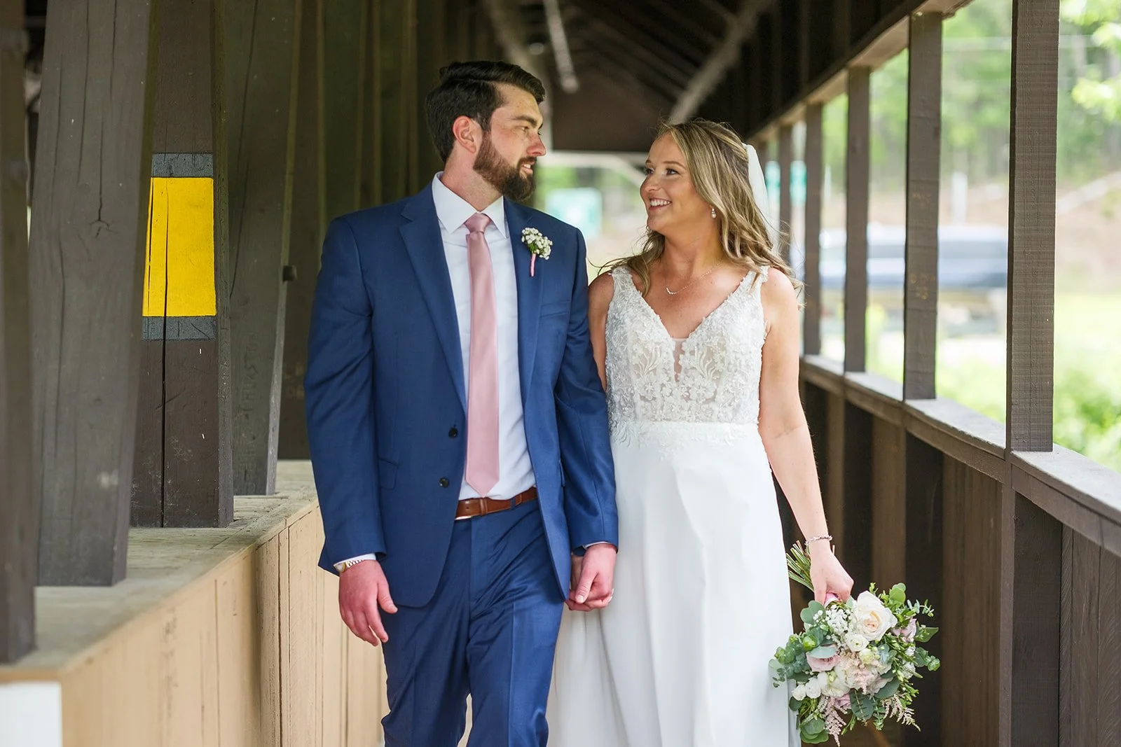 A bride and groom walk arm-in-arm on a covered wooden walkway, smiling at each other, with the bride holding a bouquet of flowers. The groom is wearing a blue suit with a pink tie, and the bride is in a lace wedding gown with a bouquet of roses and g