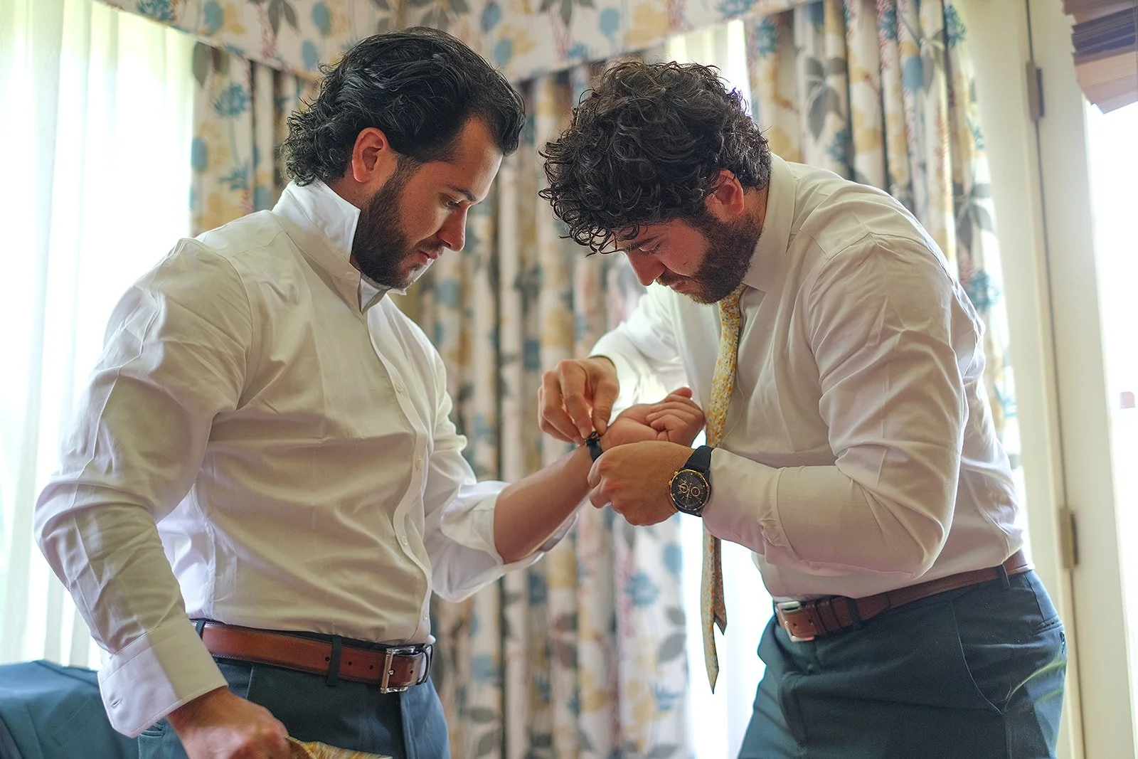 Two men in white shirts and dress pants are adjusting a watch on the wrist of the man on the right in a room with floral and striped curtains.
