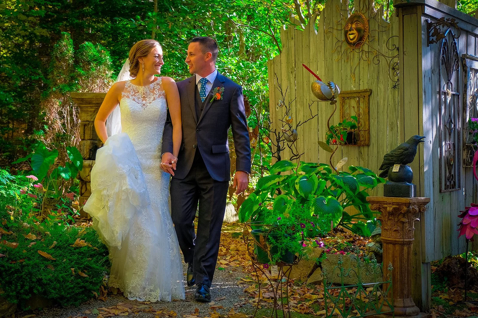 A bride and groom holding hands and smiling at each other in a garden setting surrounded by leafy green plants and garden decorations.