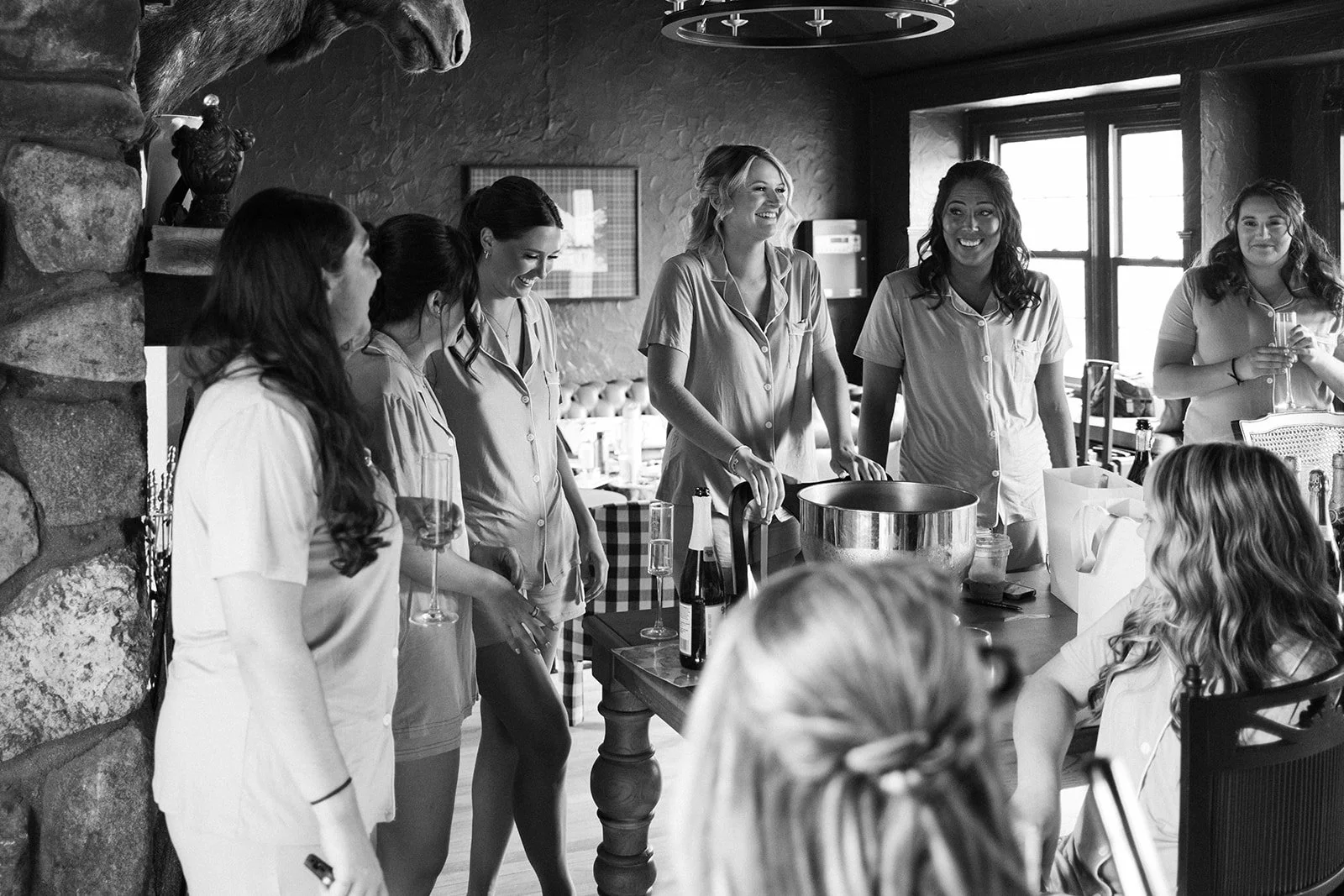 A group of women in pajamas gathered around a table, celebrating with drinks, in a cozy indoor setting with stone wall features and windows, during a bridal shower or similar celebration.