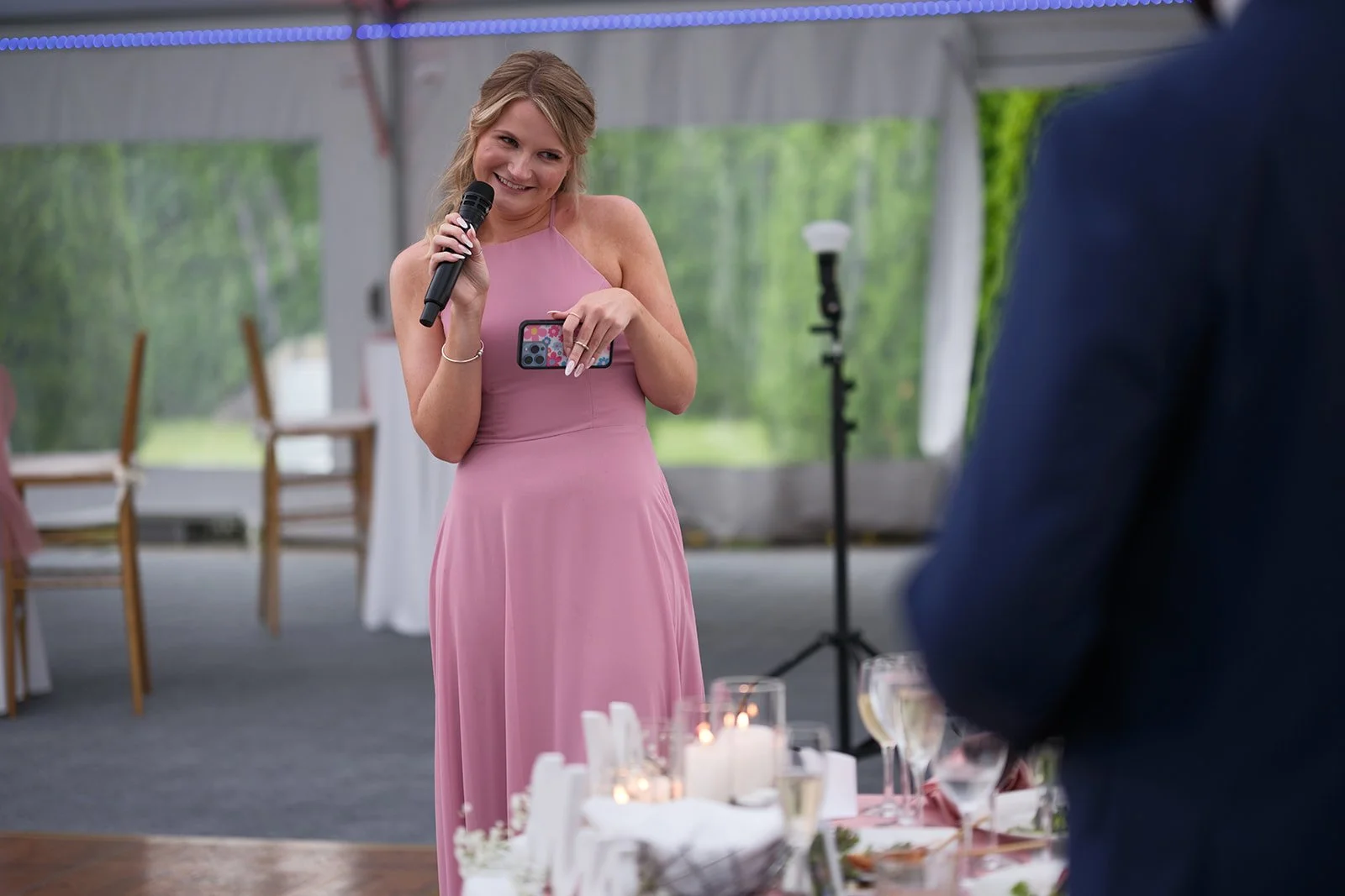 A woman in a pink dress holding a microphone and a phone, smiling at a wedding reception under a tent, with tables, glasses, candles, and a man in a dark suit nearby.