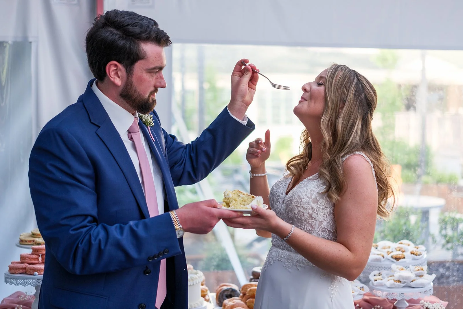 A groom feeding a slice of cake to a bride at their wedding reception, with desserts visible in the background.