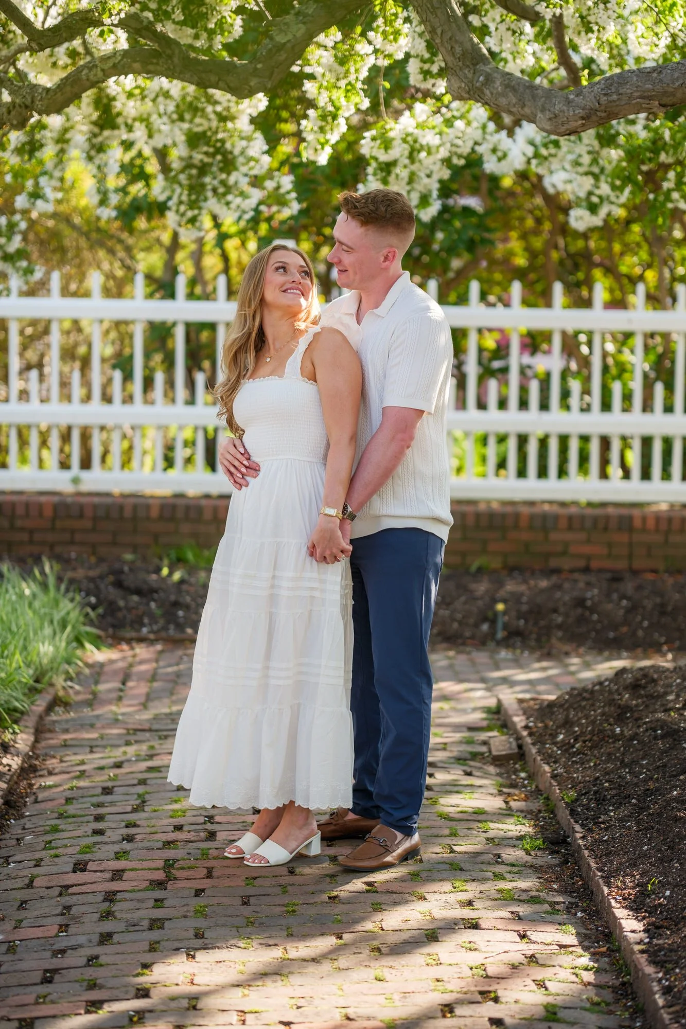 Engaged couple Melanie and Connor hold hands and gaze at each other on a brick garden path beneath a blooming tree at Prescott Park in Portsmouth, NH