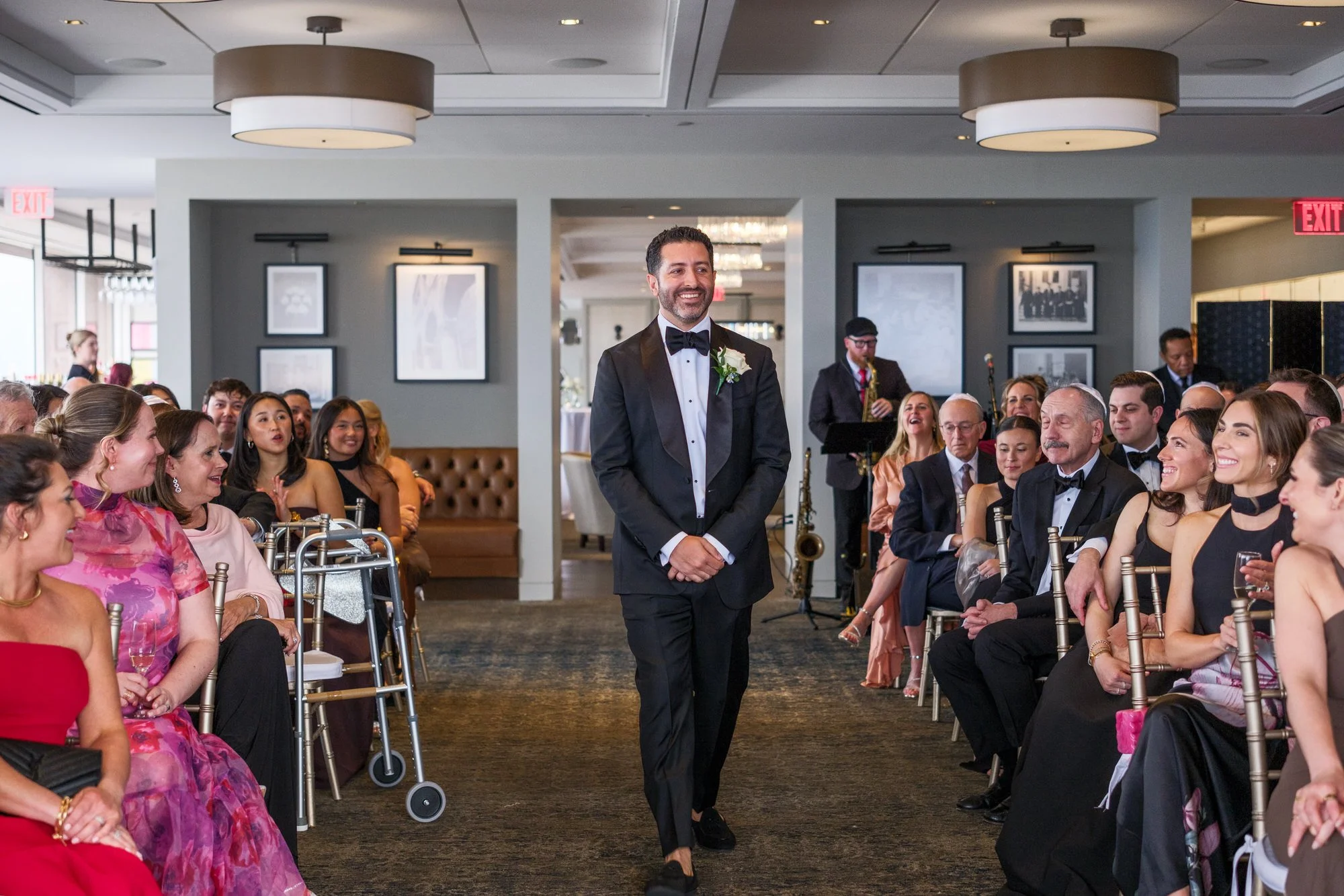 Groom walking down the aisle during his wedding processional at the UMass Club Boston with live saxophonist and guests looking on