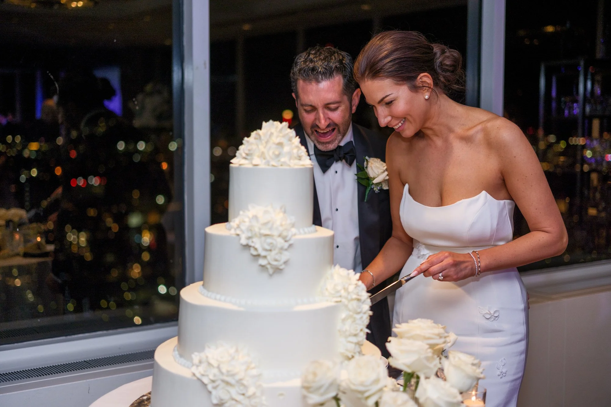Bride and groom cutting their elegant white tiered wedding cake with Boston city lights glittering behind them