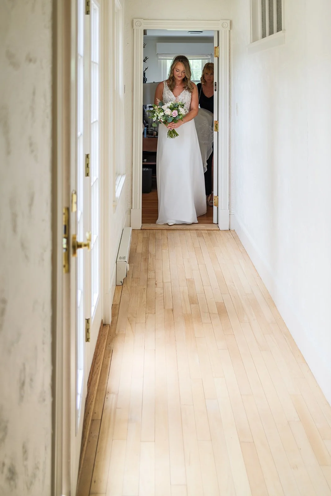 A bride in a white wedding dress holding a bouquet of flowers walking through a doorway, with a woman following behind her, in a well-lit hallway.