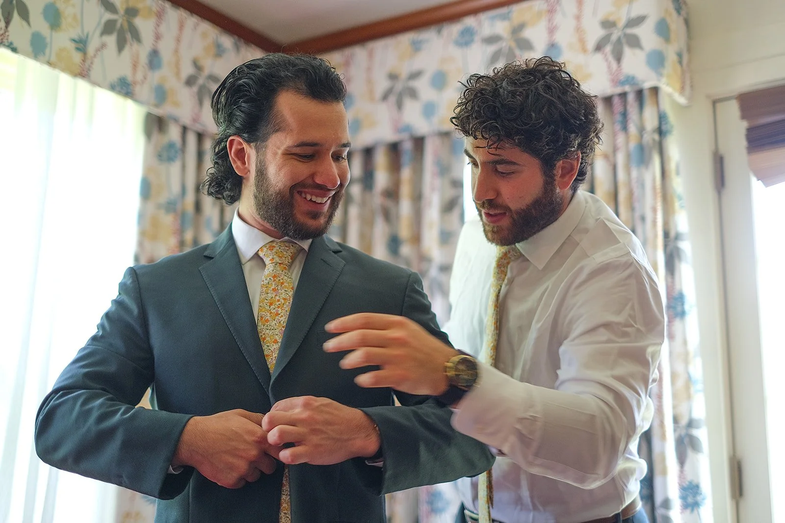 Two men, dressed in formal attire with matching floral ties, are smiling and interacting in a room with floral curtains and natural light.