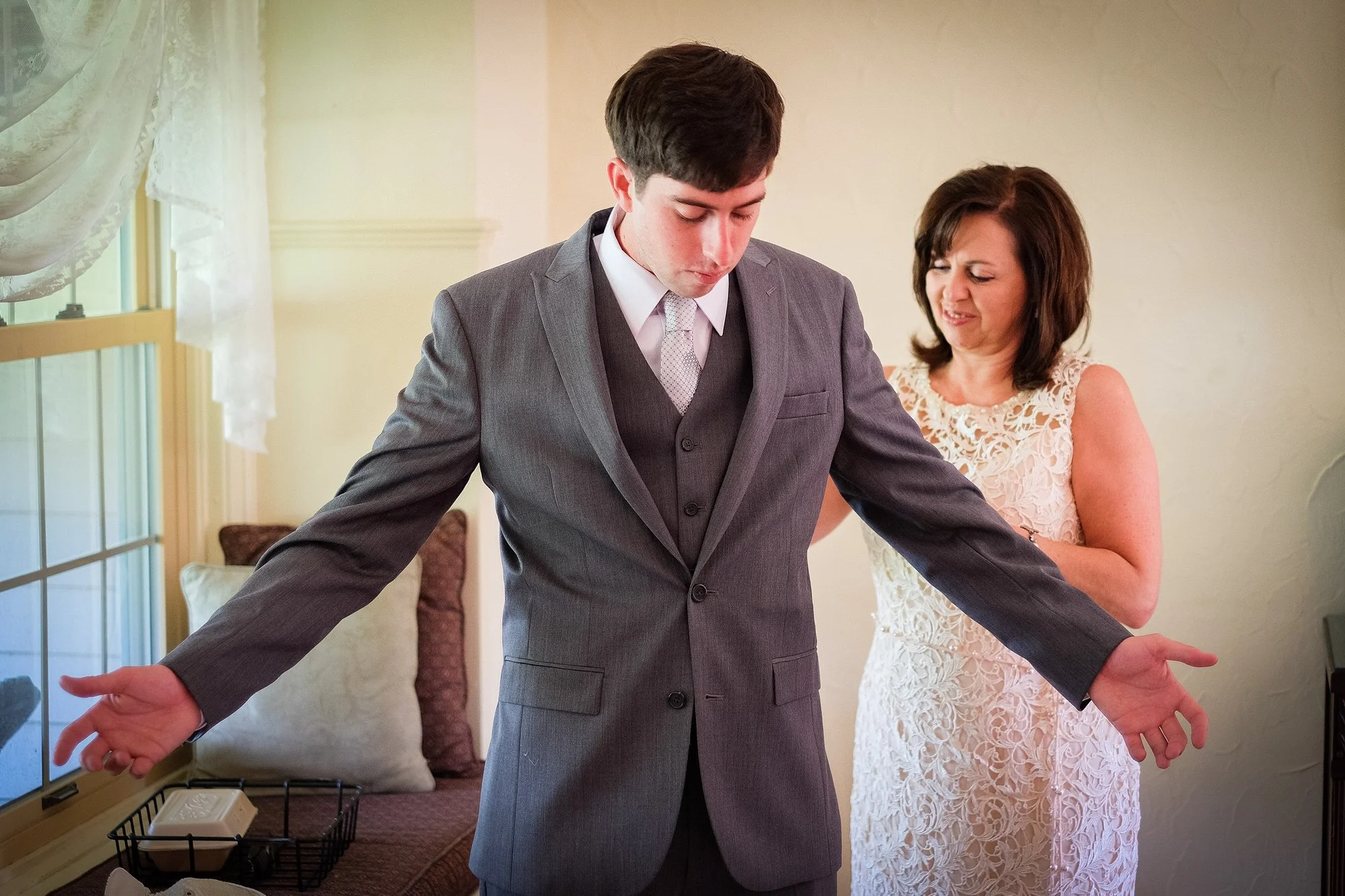 A young man in a gray suit and tie stands with his arms outstretched, looking down, while a woman in a cream lace dress stands behind him, appearing to be adjusting or fixing his suit.