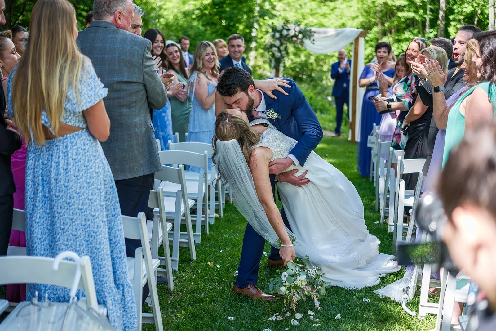 A groom dips and kisses a bride during an outdoor wedding ceremony, with guests clapping and smiling around them.