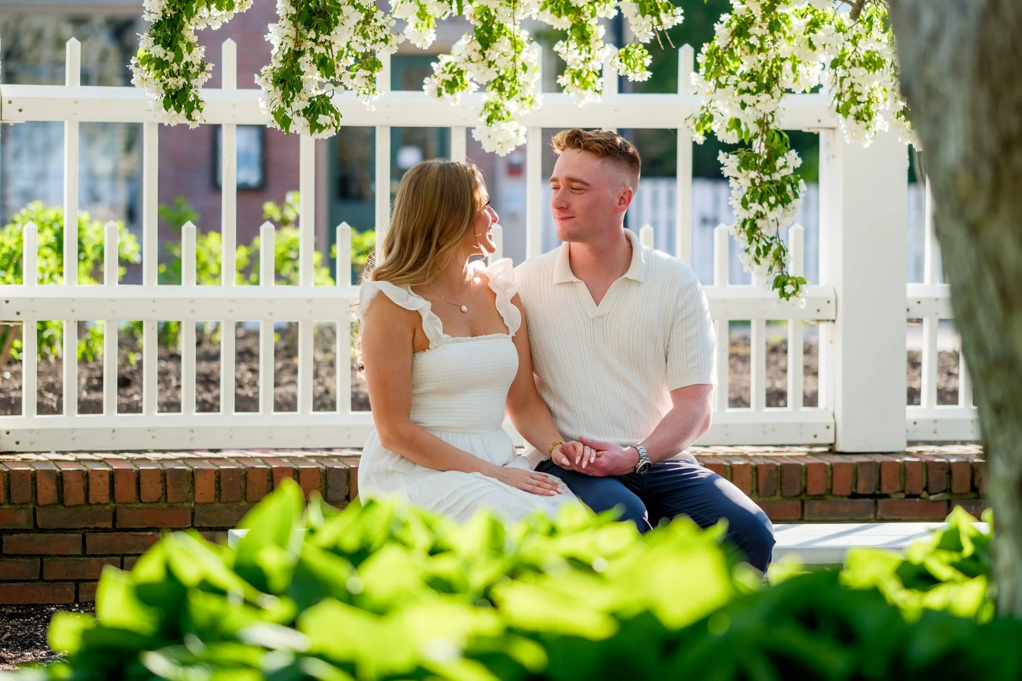 Melanie and Connor walk hand-in-hand down a brick garden path lined with white flowering trees at Prescott Park, Portsmouth, NH