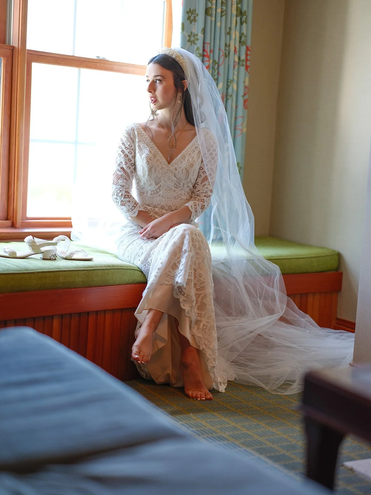 A bride sitting on a window seat in a room, wearing a lace wedding dress and veil, looking out the window with shoes placed beside her.