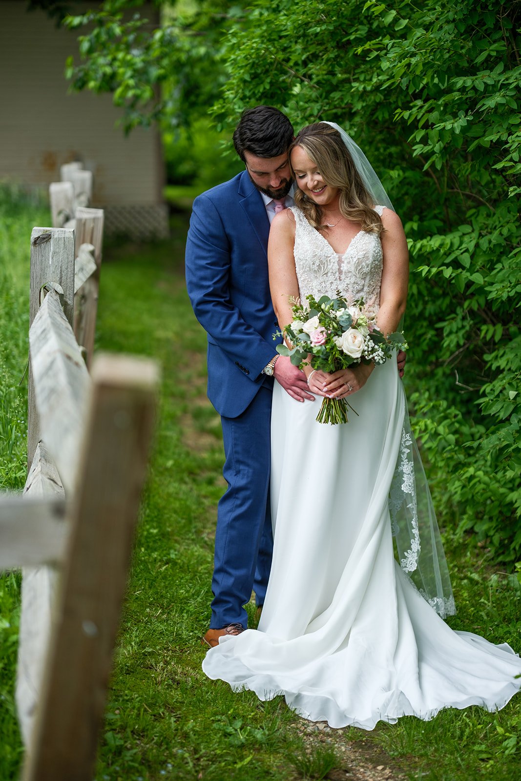 Bride and groom standing on a grassy pathway with green bushes in the background, the groom is holding the bride from behind as they smile. The bride wears a white wedding dress with lace details and a veil, holding a bouquet of white and pink roses 