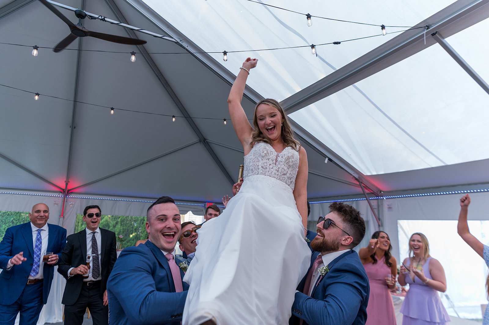A bride being lifted on a wedding reception dance floor, smiling and raising her arm with friends and family around her, under a tent with string lights.