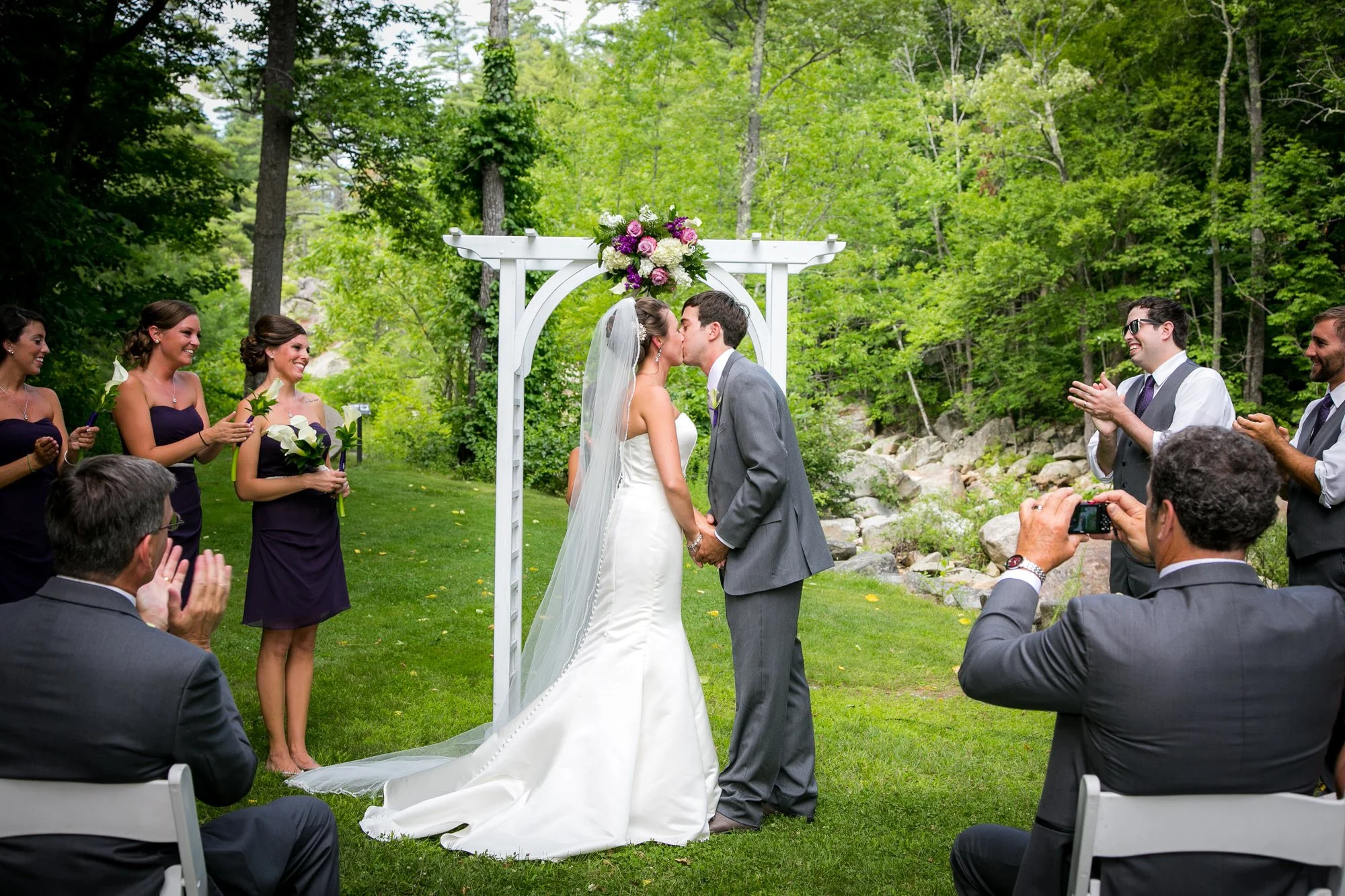 A bride and groom share a kiss during an outdoor wedding ceremony under a white arbor decorated with flowers, surrounded by bridesmaids and groomsmen in a green, wooded area.