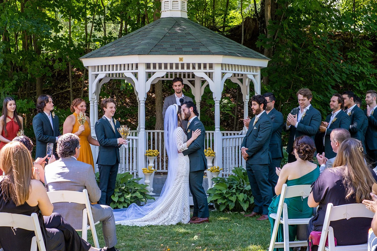 A wedding ceremony taking place outdoors in front of a small white gazebo, with a bride and groom kissing at the altar while guests watch and take photos.