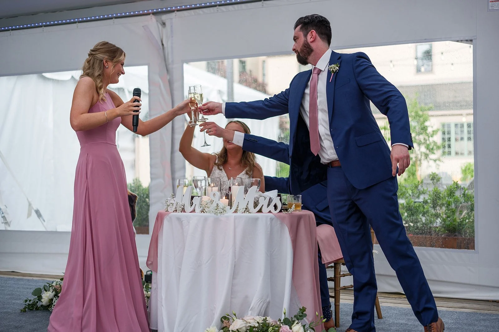 A wedding reception scene where a woman in a pink dress is giving a toast, and a man in a blue suit is toasting her with champagne, with a woman in white seated at the head table.