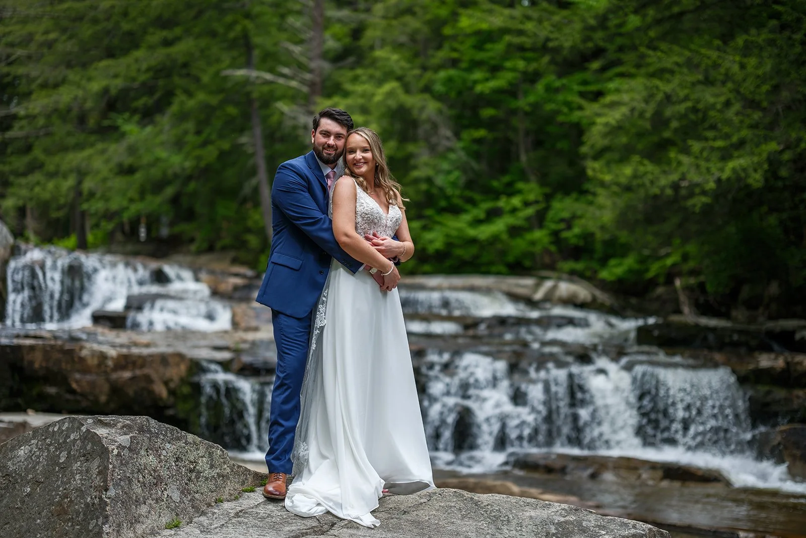 A newlywed couple standing on a rock in front of a small waterfall surrounded by lush green trees.