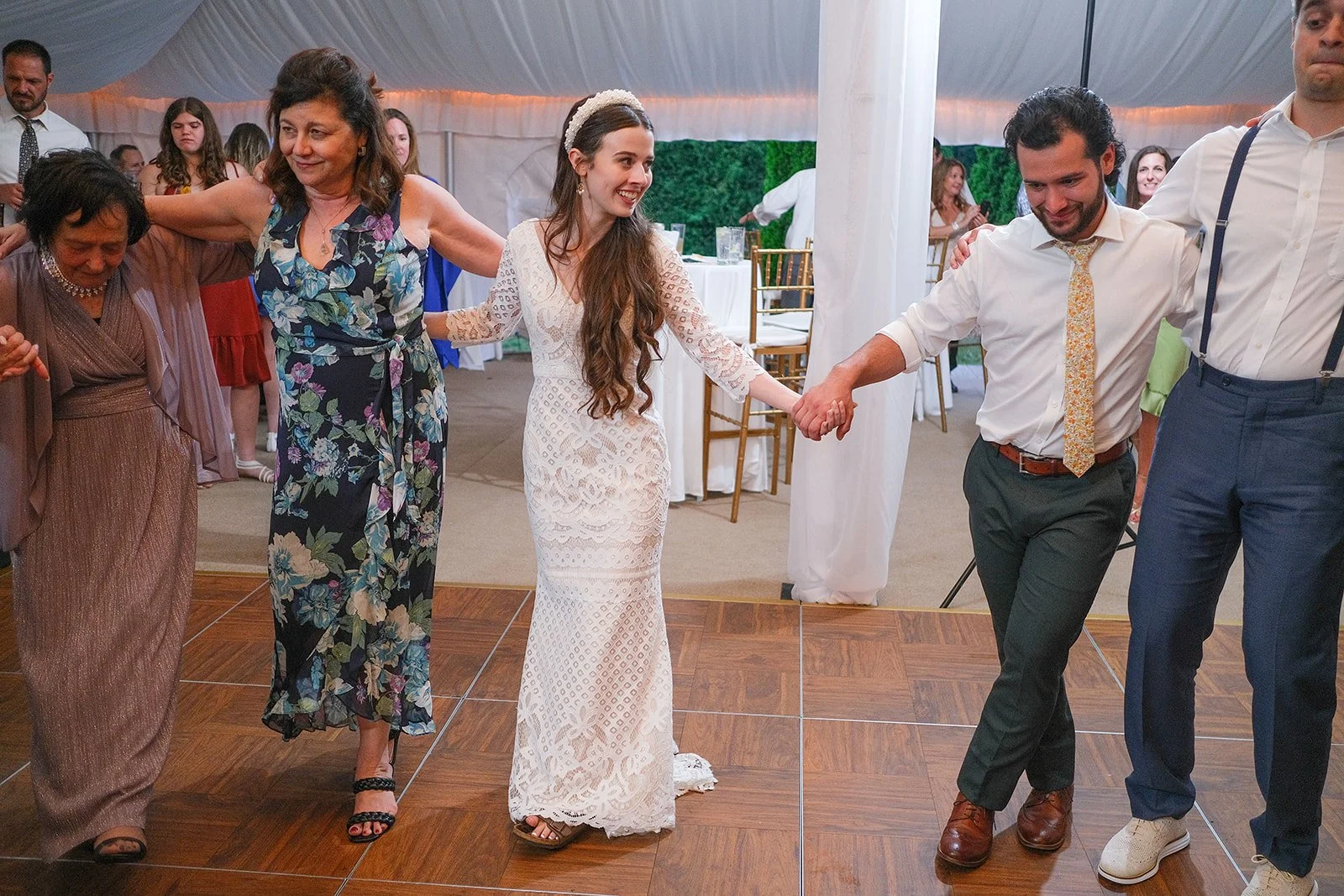 People holding hands in a dance circle at a wedding reception, smiling, with a woman in a white lace dress in the center.